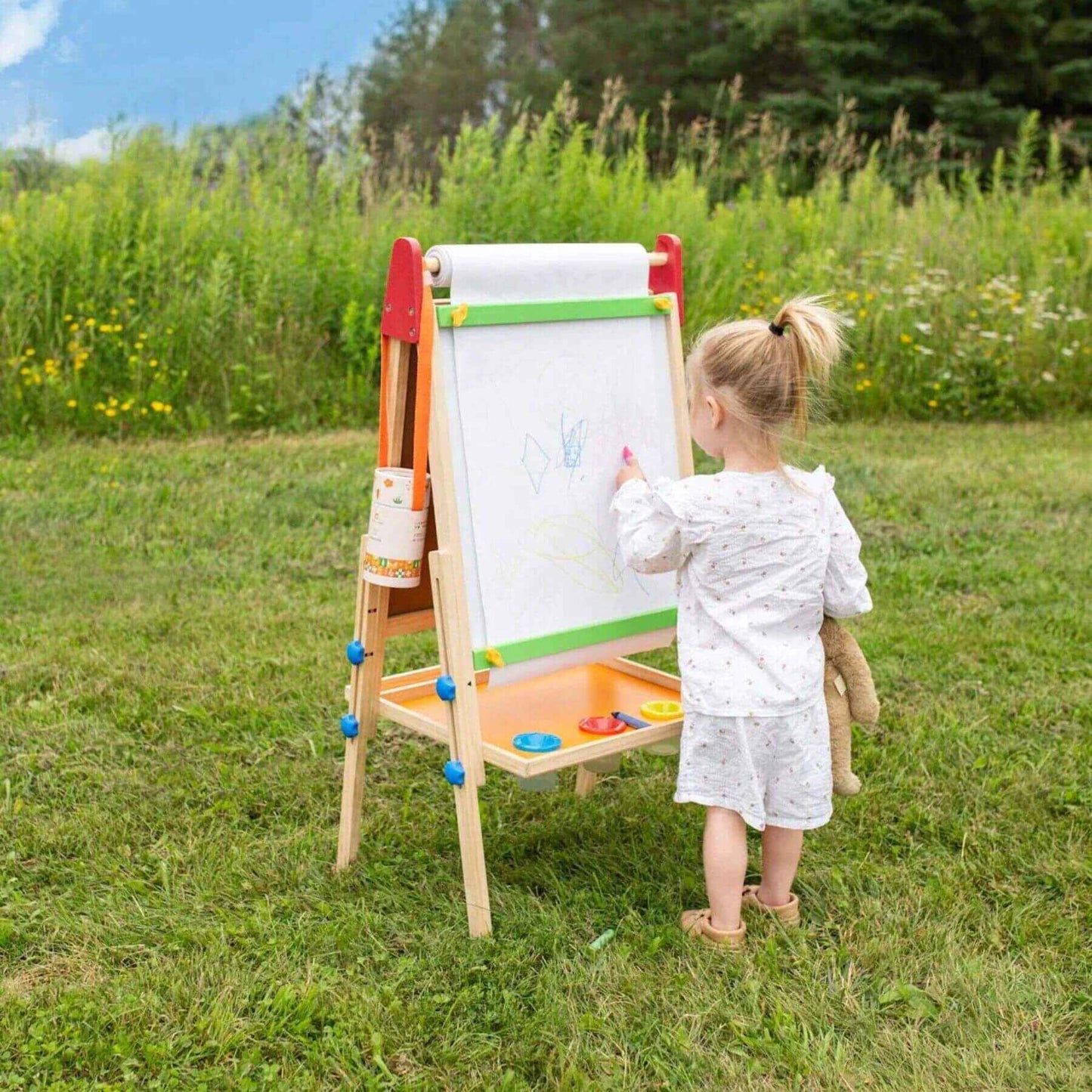 Child drawing on Tiny Land® 3-in-1 Art Easel & Paper Roll Set in a grassy area with trees and flowers in the background