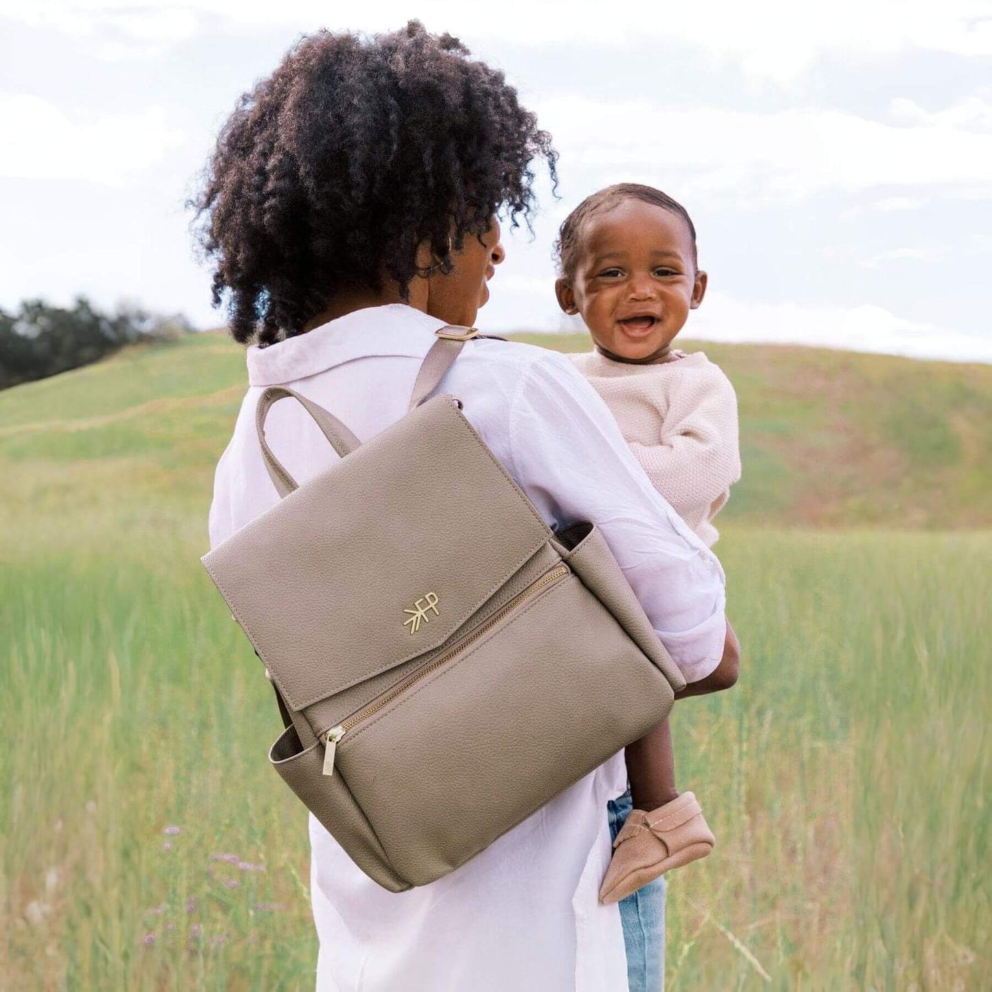 Woman carrying a child on her back with Freshly Picked Aspen Mini Classic Bag II in a grassy field