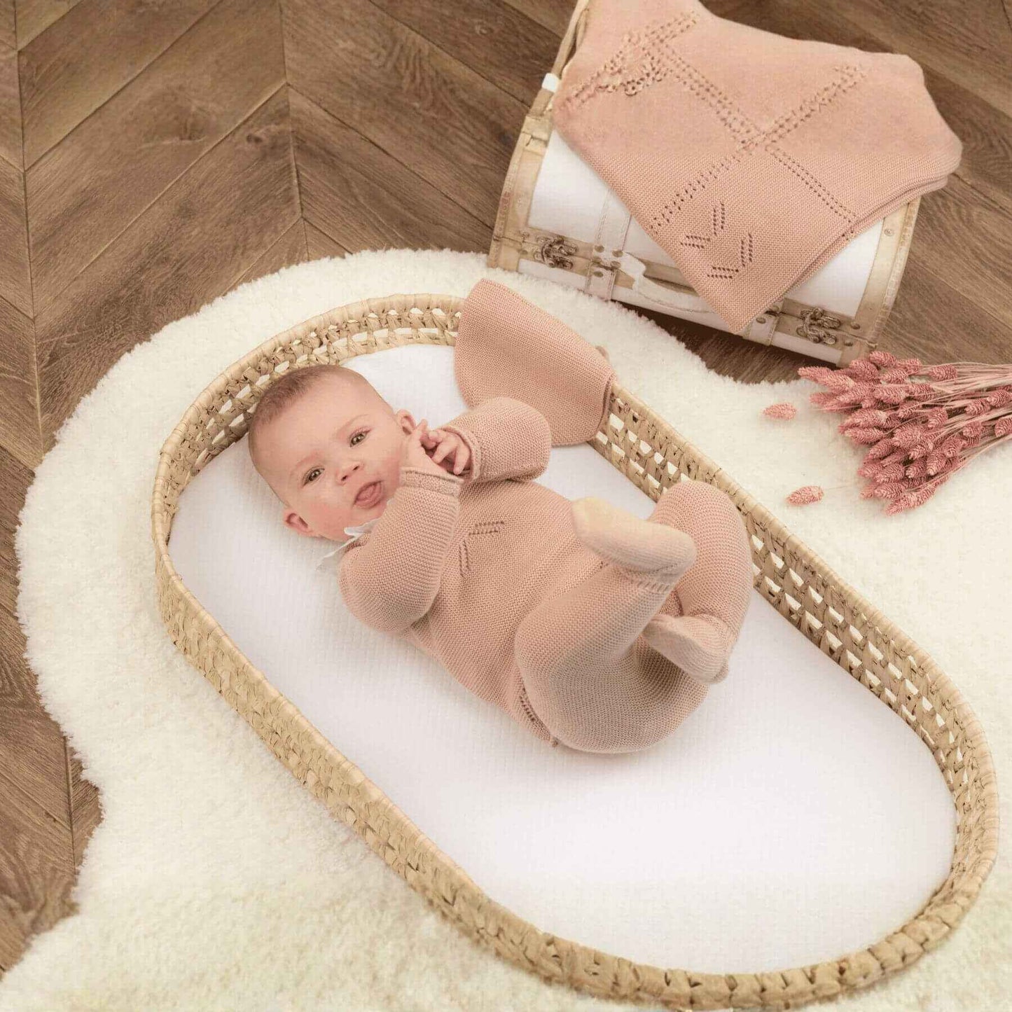 Baby in a wicker basket on a wooden floor with a pink blanket and flowers.