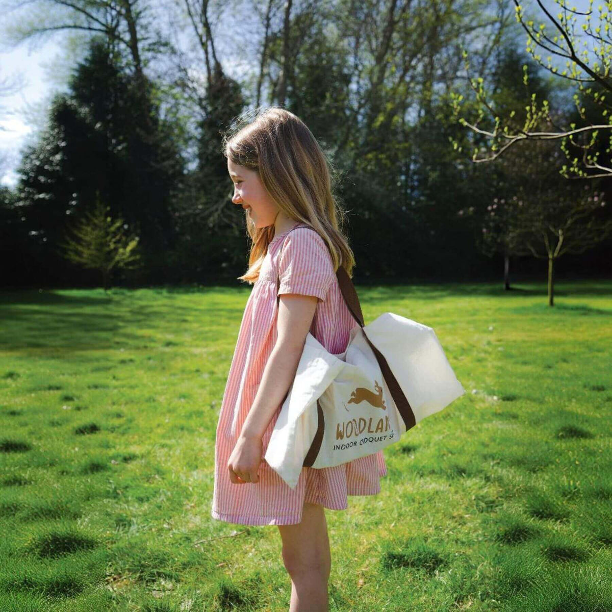 Young girl in a pink dress holding a white tote bag with a logo in a grassy field.