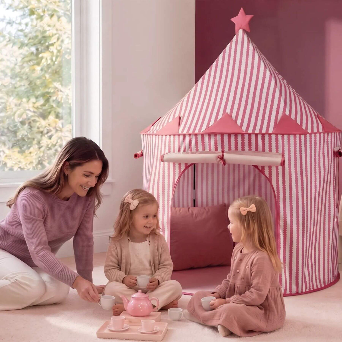 A woman and two young girls are gathered in a playroom with Tiny Land® Kids Tent - Pink. The woman is kneeling and pouring tea from a pink teapot on a tray with teacups, while the girls sit inside the tent and around her.