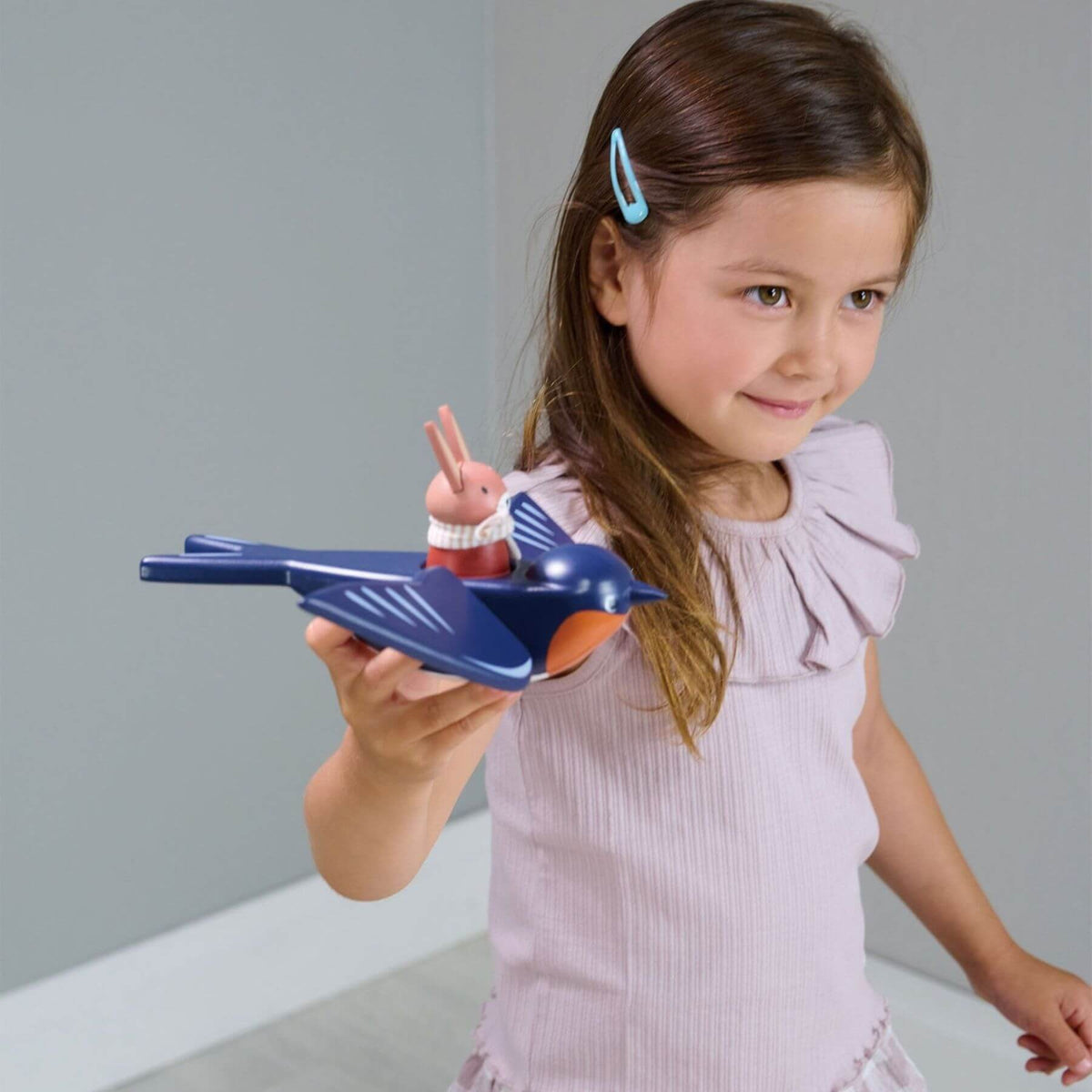 A young girl with brown hair and a light blue hair clip holds a blue bird-shaped toy bird with a small rabbit figure on top, set against a neutral gray background.