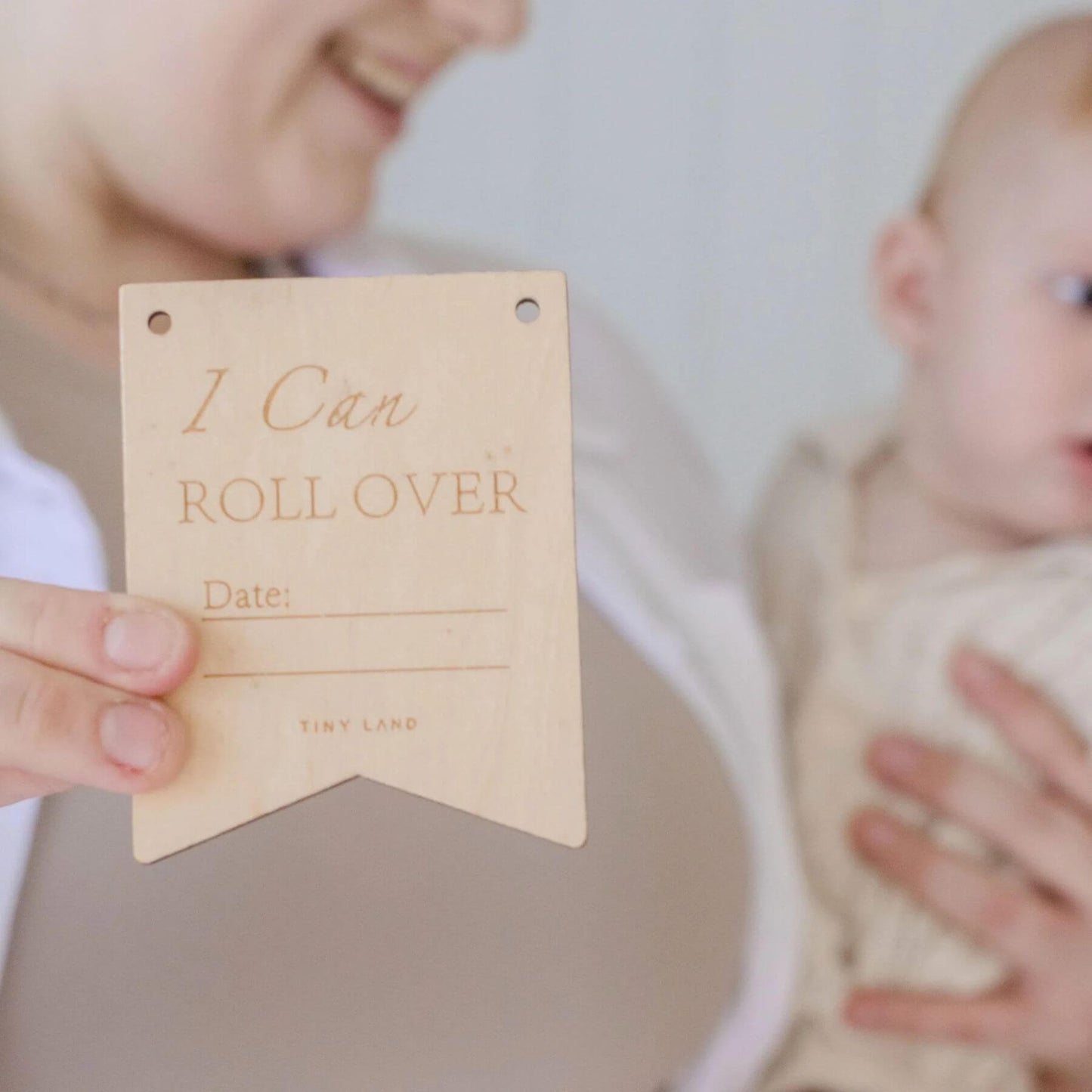 Person holding a wooden milestone tag with 'I Can Roll Over' text, blurred background