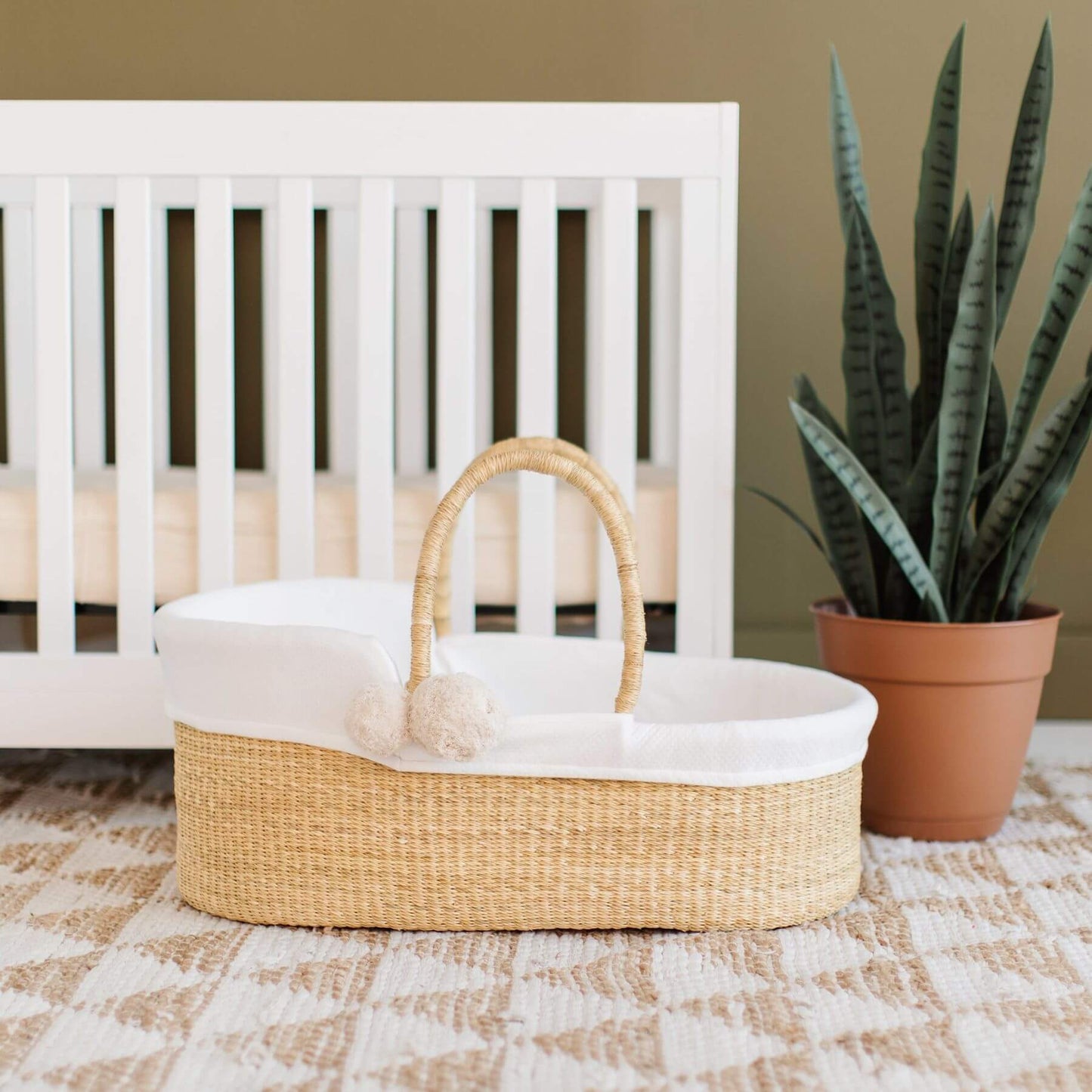 Plum+Sparrow African Moses Basket Vegan Handle in front of a white crib with a potted plant on a patterned rug.