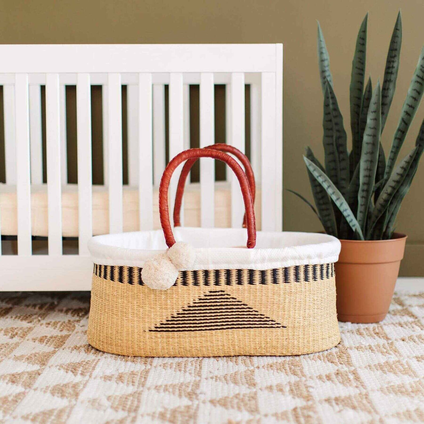 Plum+Sparrow Artisan Sky No Hood African Moses Basket on a checkered floor next to a white crib and potted plant.
