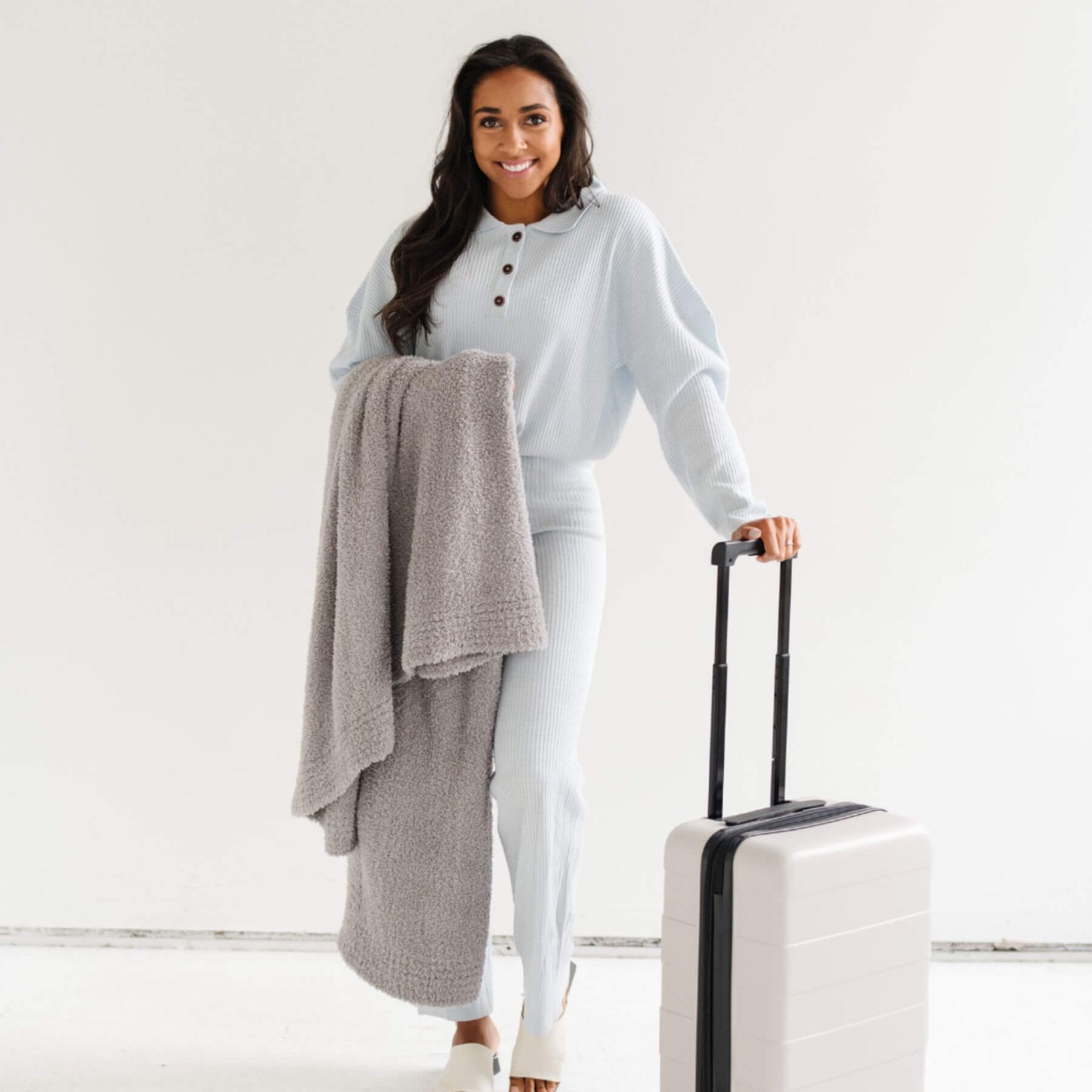 Woman holding Saranoni Bamboni® Throw Blanket, and a suitcase with a light gray outfit on a white background