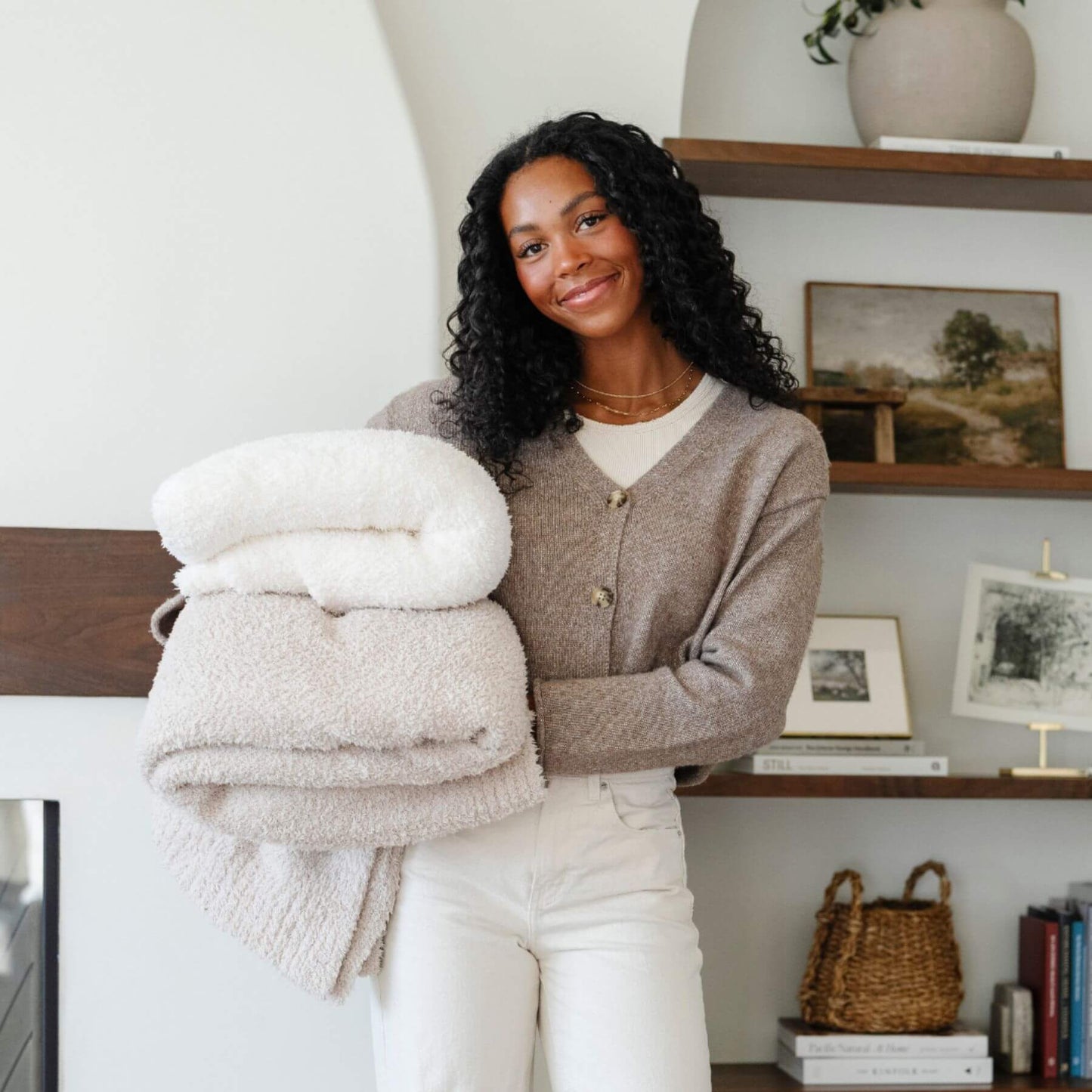 Woman holding a stack of folded Saranoni Bamboni® Throw Blankets in a cozy room with shelves and decor.