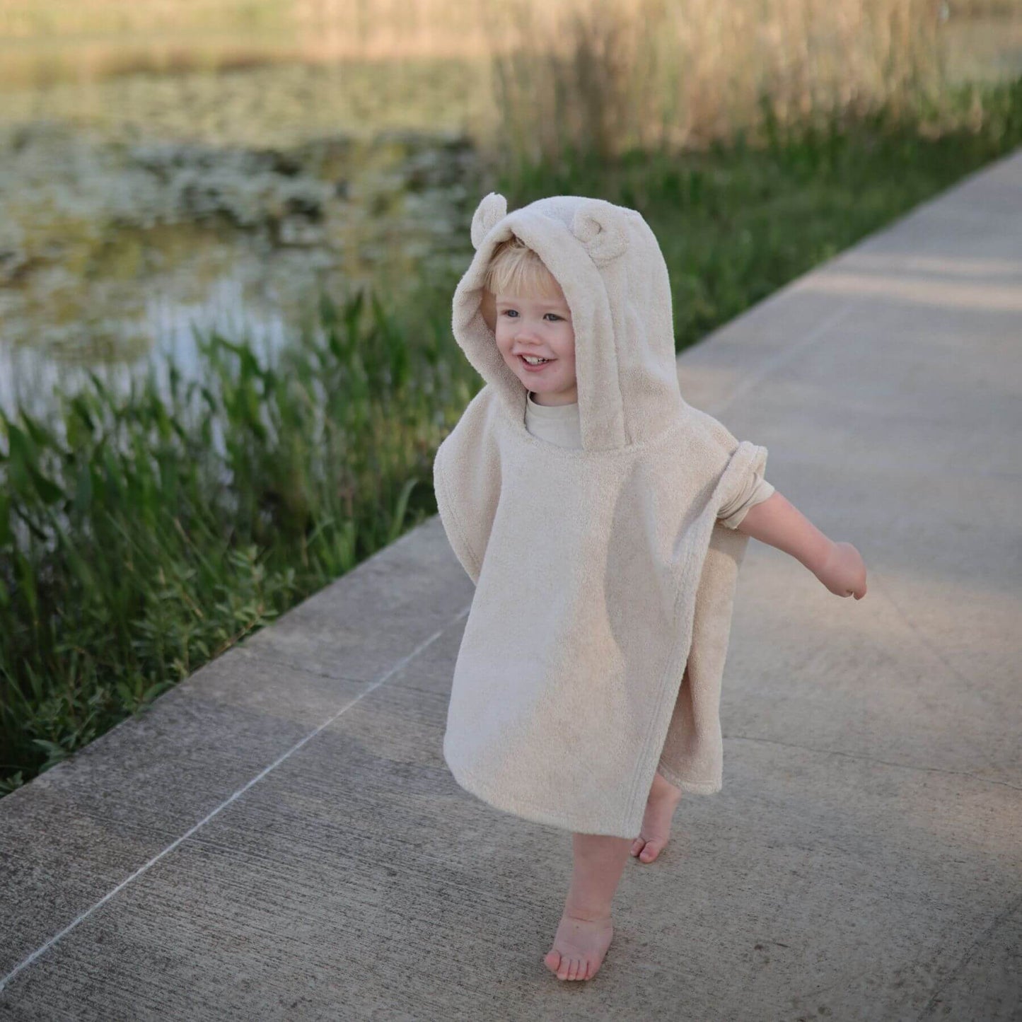 Child wearing a Mushie Bear Poncho Towel outdoors by a body of water