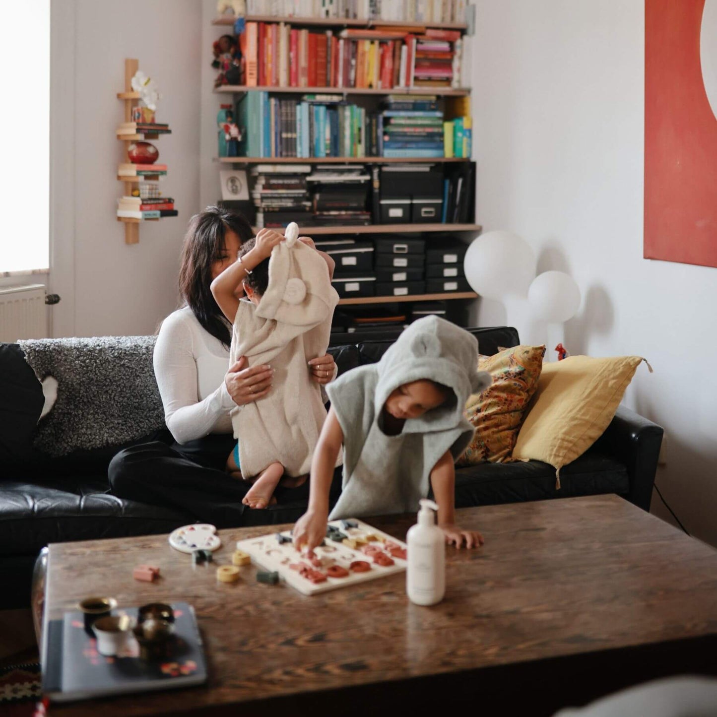 Two children in Mushie Bear Poncho Towel playing with food on a table in a living room.
