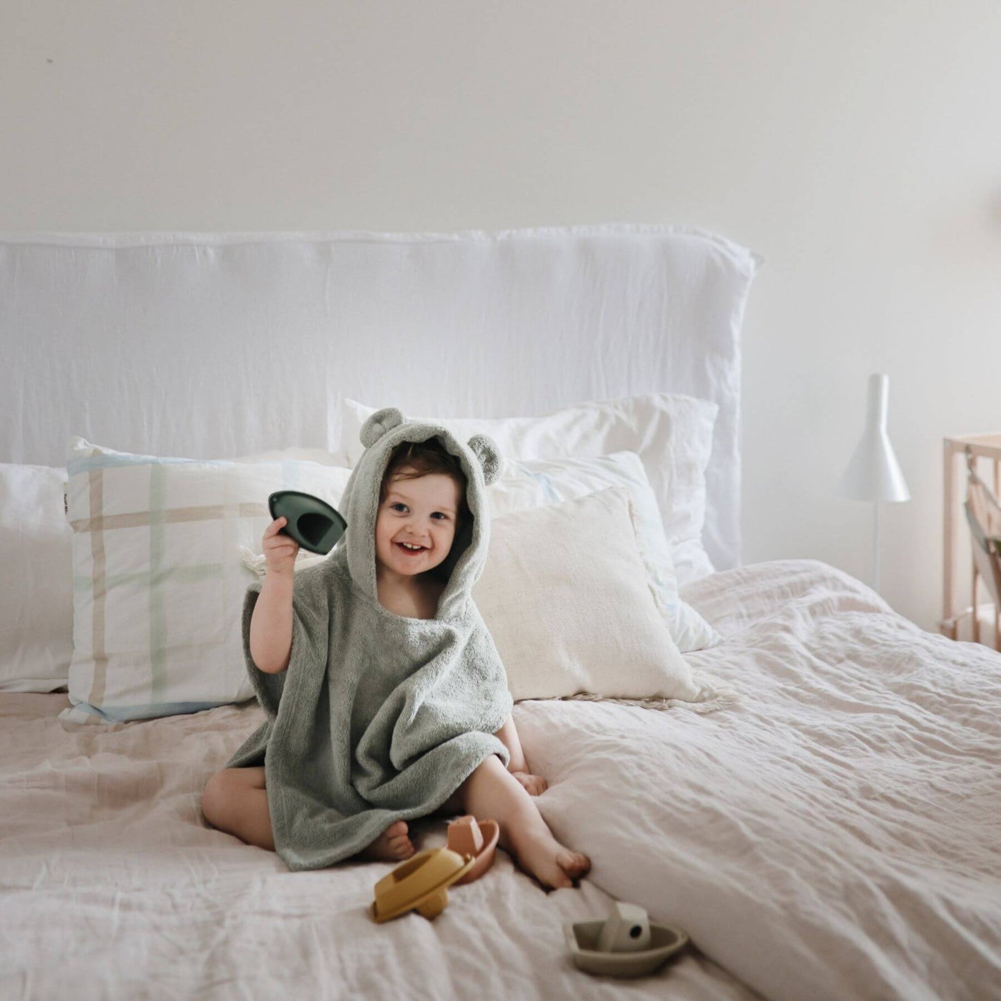 Child in Mushie Bear Poncho Towel sitting on a bed holding a toy.