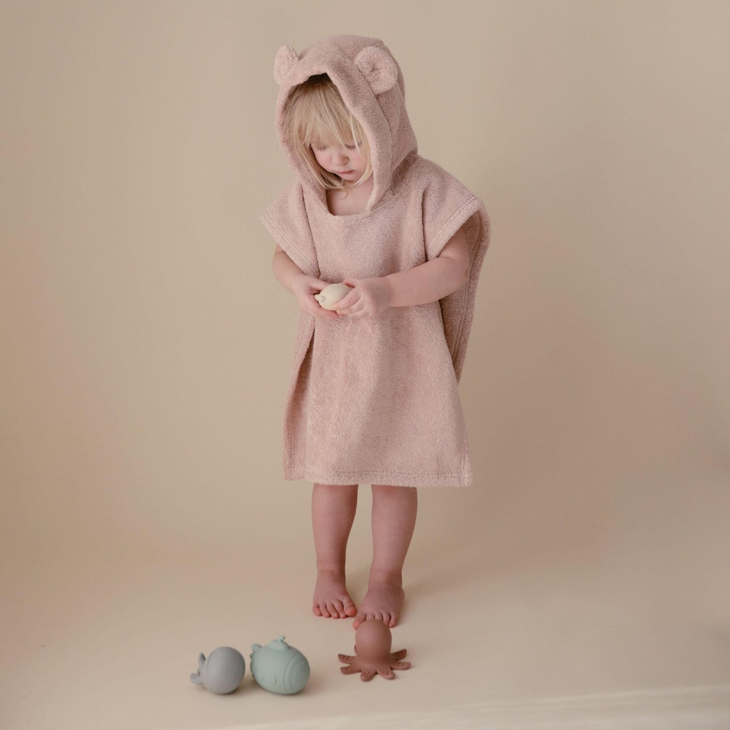 Child wearing Mushie Bear Poncho Towel with bear ears, standing on a beige background.