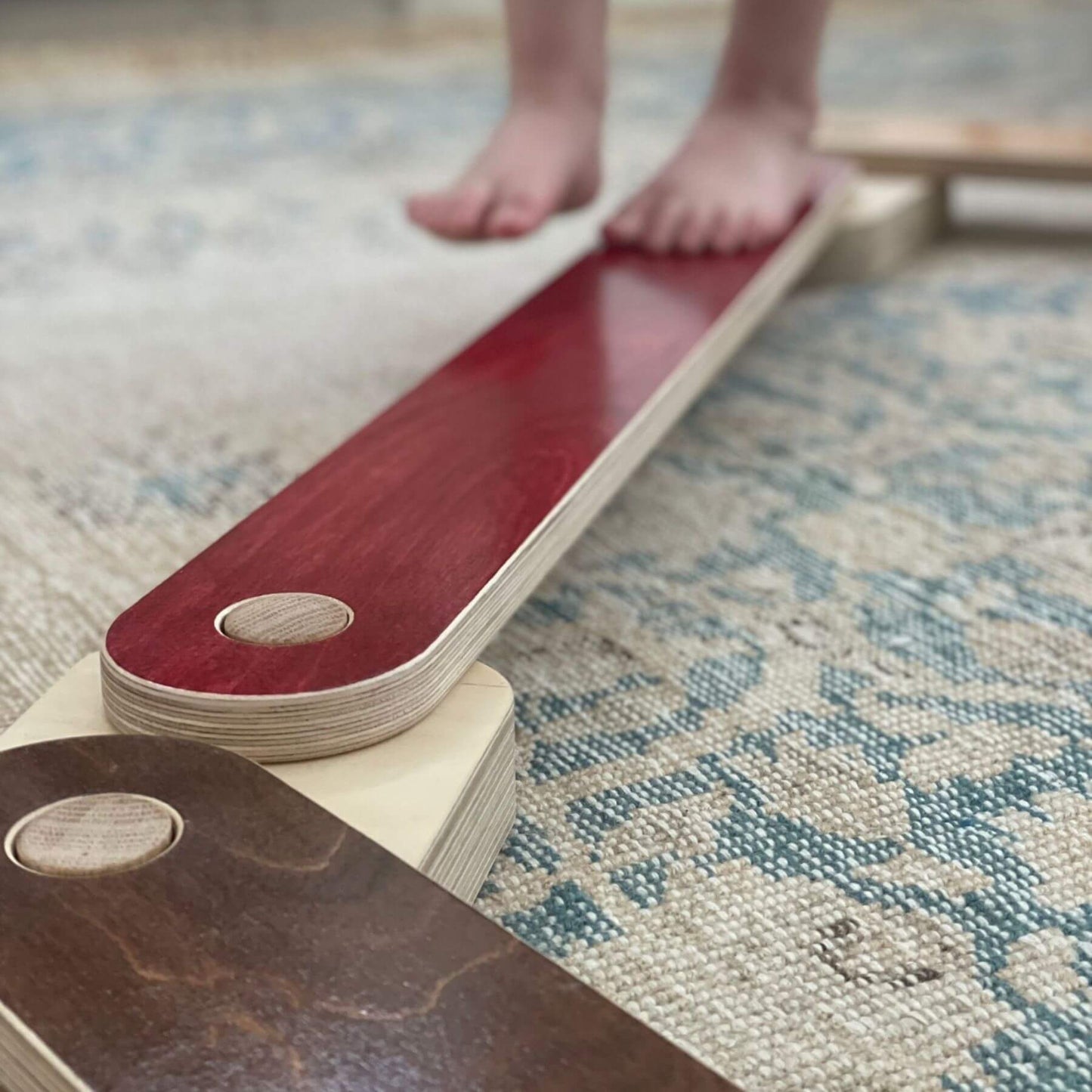 Wooden balance board on a patterned rug with a child's feet standing on it.