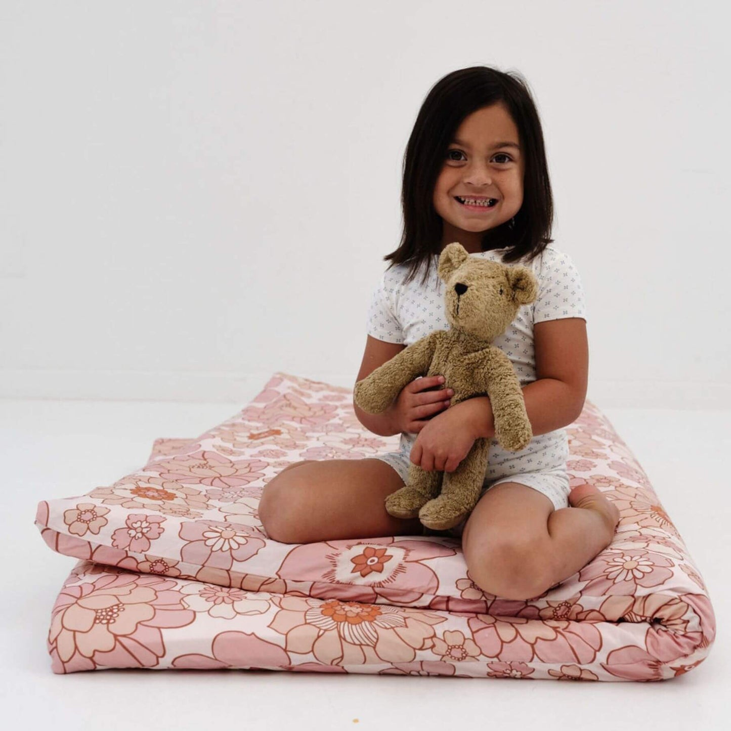 Child sitting on folded Toki Mats Blooms Organic Cotton Mat holding a teddy bear against a white background