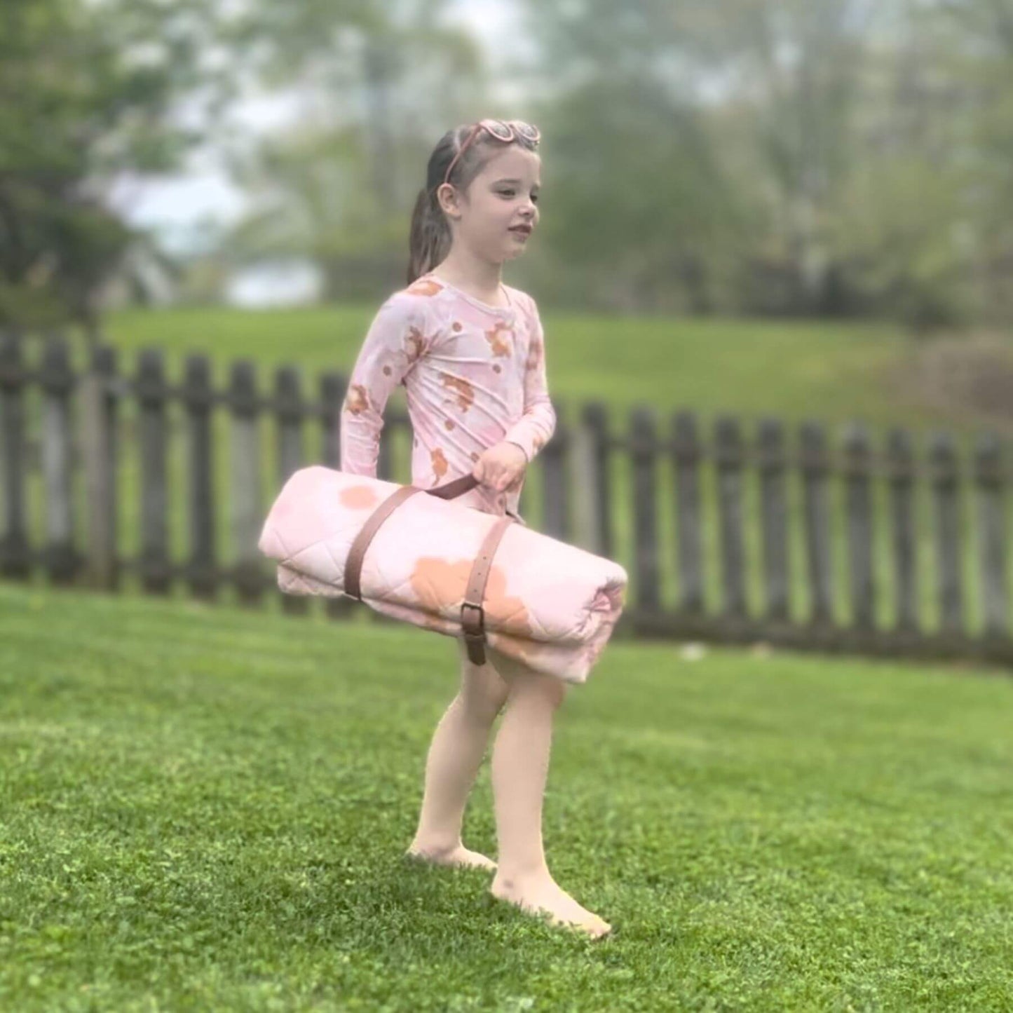 Young girl holding Blush Meadow Quilted Water Resistant Beach Mat on grass with a wooden fence in the background