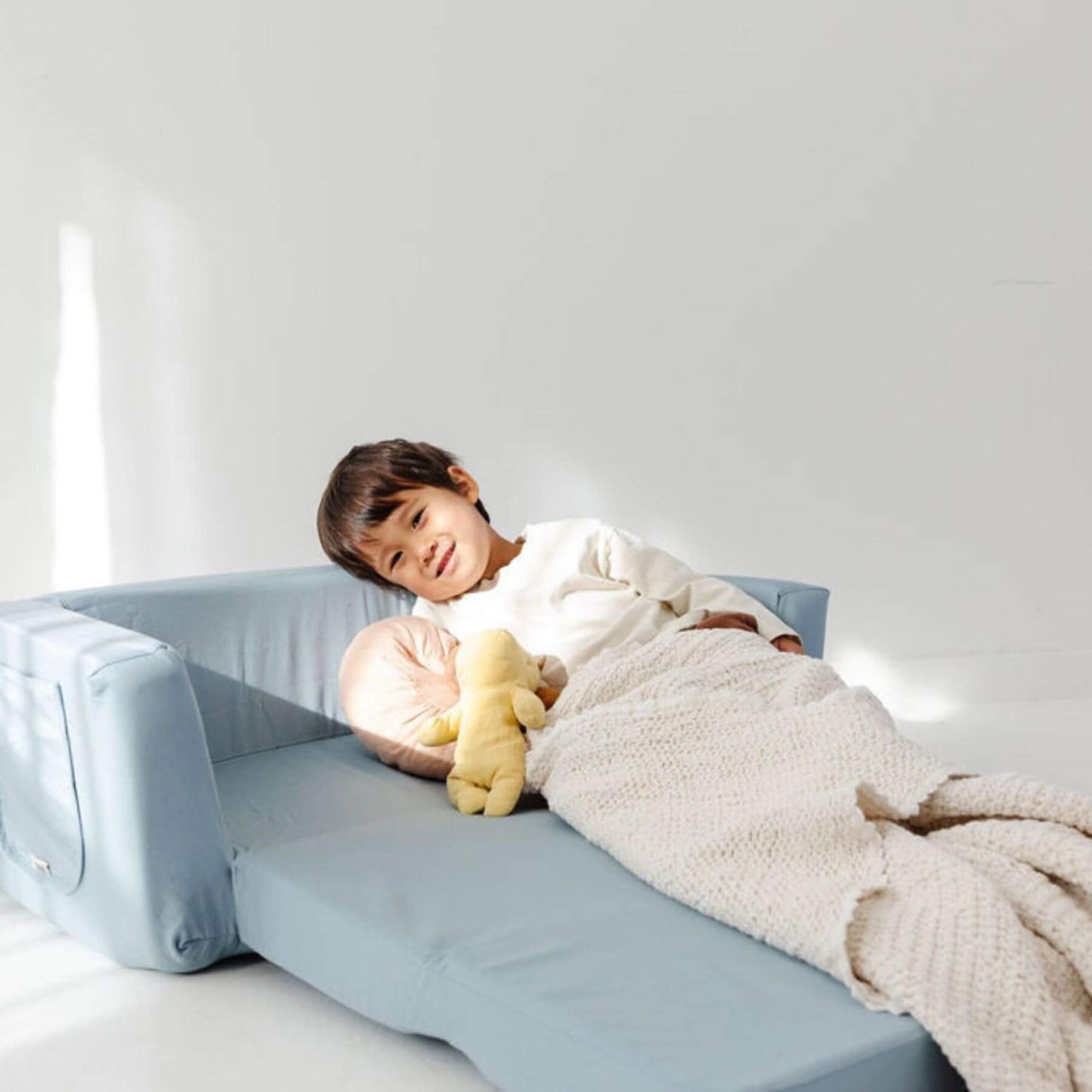 Child lying on Toki Kids Breeze Play Couch with a white blanket and yellow toy, smiling.