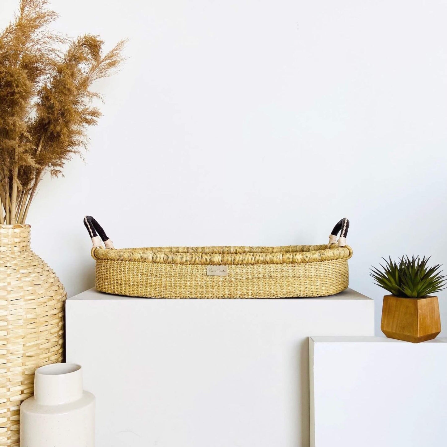 Decorative setup with Plum+Sparrow Changing Basket Brown+Cream Handle, vase, and plant on a white surface.