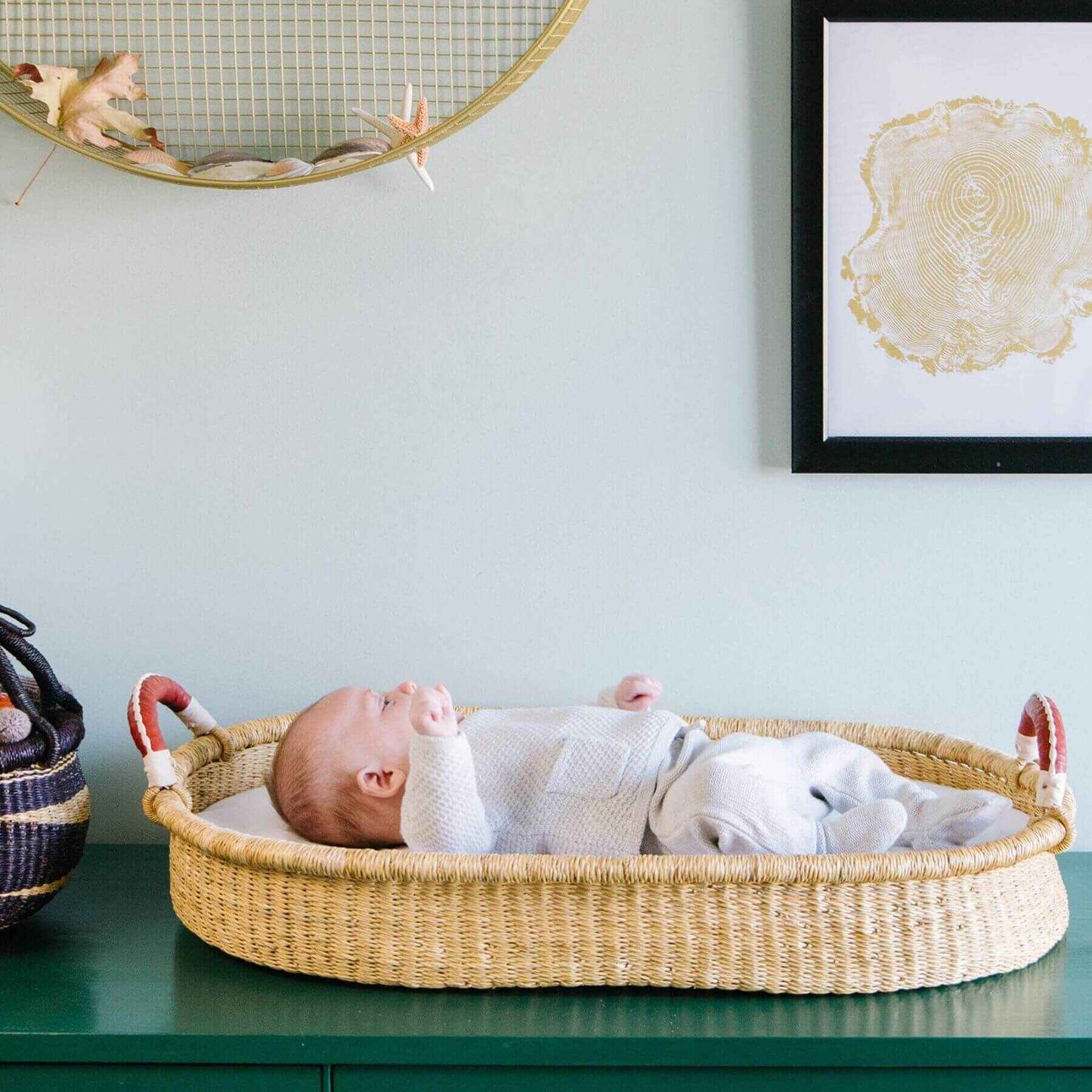 Baby lying in Plum+Sparrow Changing Basket Brown+Cream Handle on a green surface with a light blue wall and decorative items.