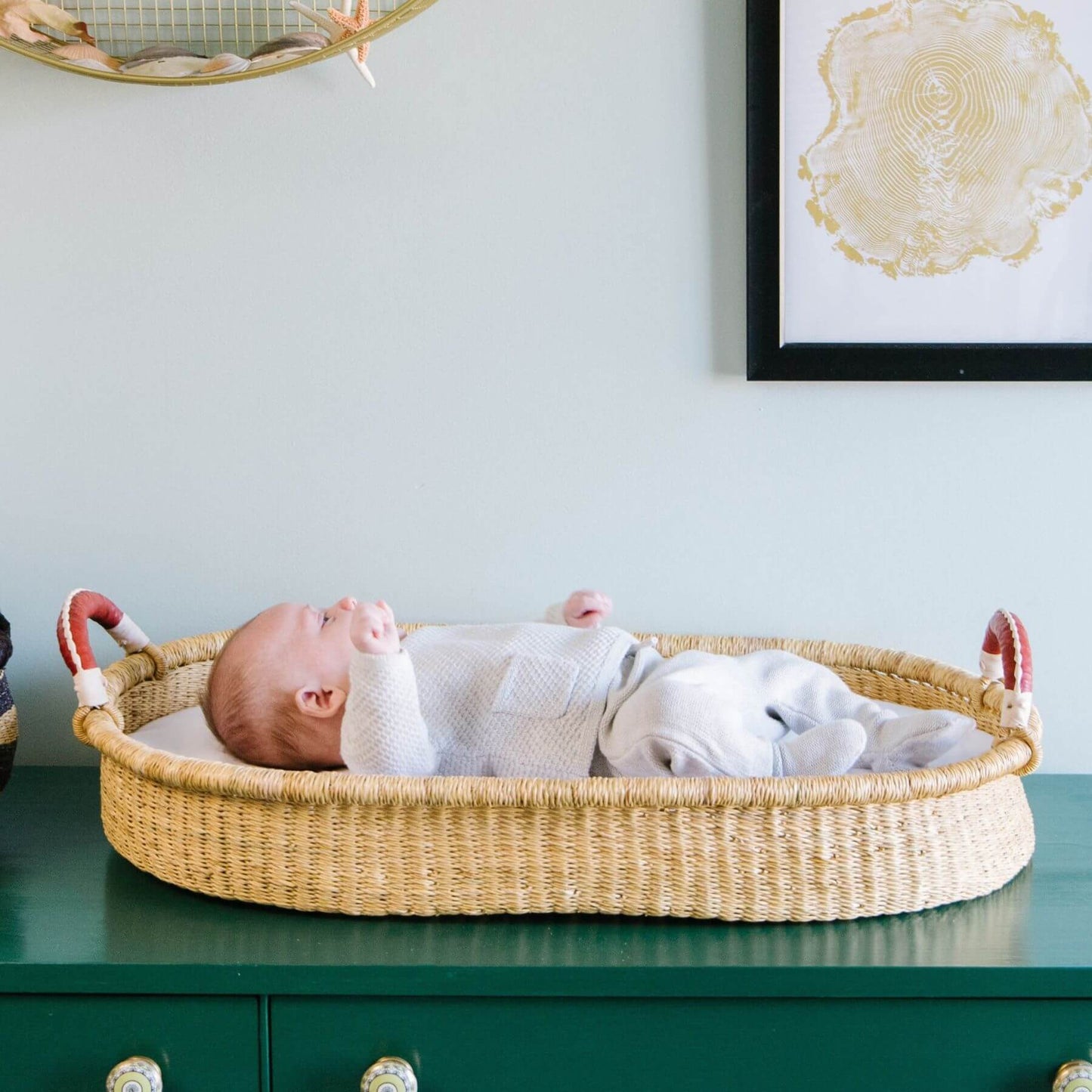 Baby lying in Plum+Sparrow Changing Basket Cognac + Cream Handle against a light blue wall with a framed picture.