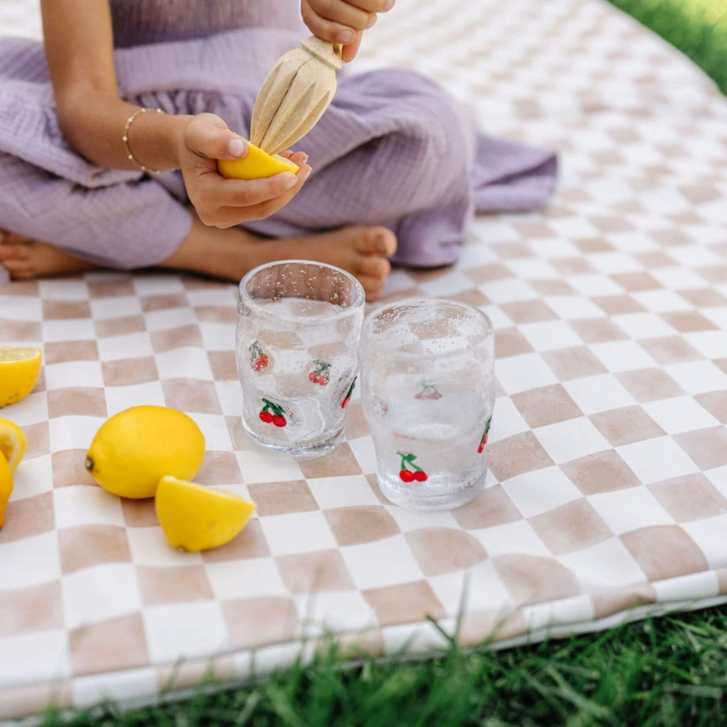 Person squeezing a lemon over two glasses on Toki Kids Checker Toffee Vegan Leather Mat outdoors