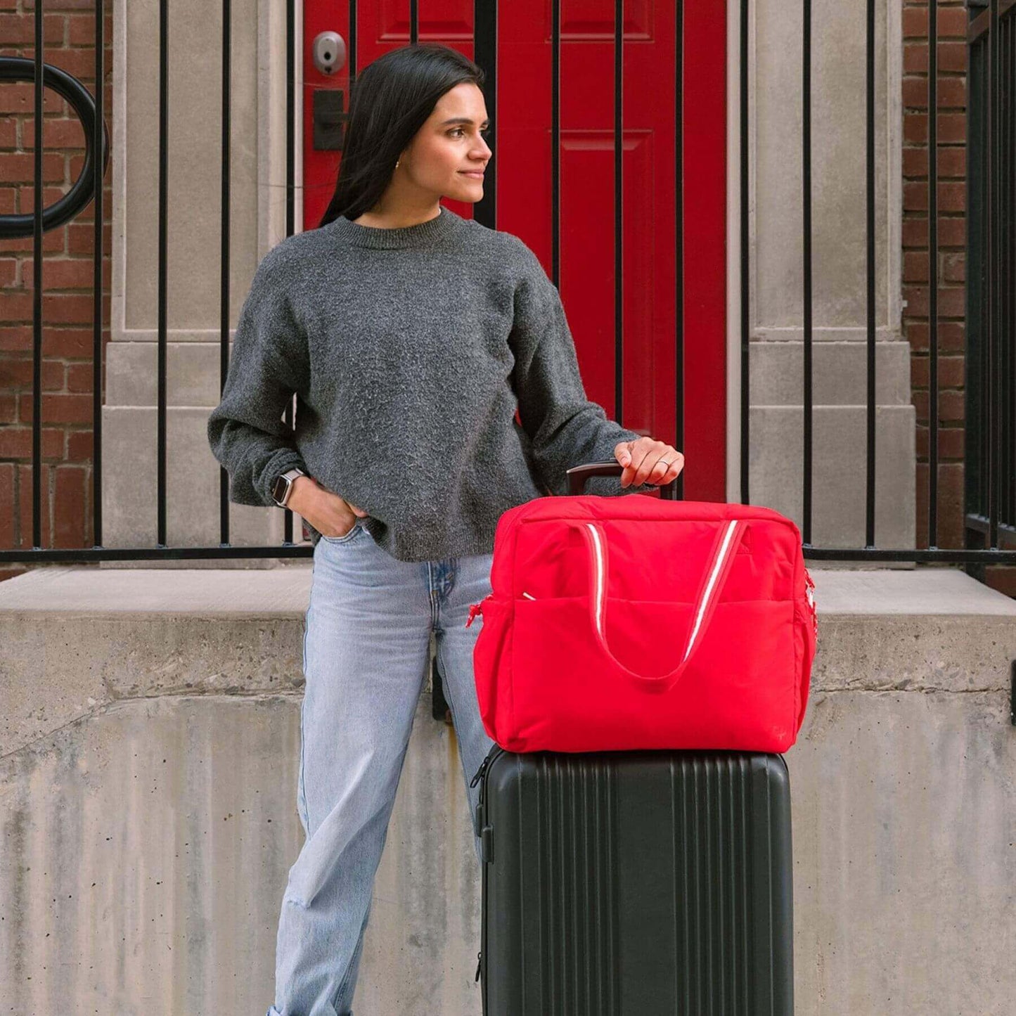 Woman holding Cherry Motion Overnight Duffle over a black suitcase in front of a red door.