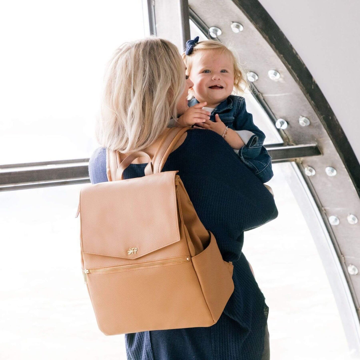 Woman holding a child wearing Butterscotch Classic Diaper Bag II in an indoor setting