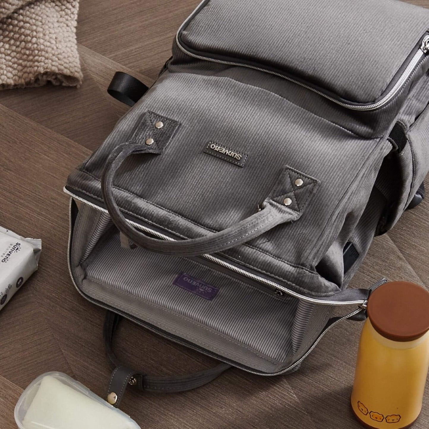 Corduroy Diaper Backpack Grey on a wooden floor with a yellow thermos and white container nearby.