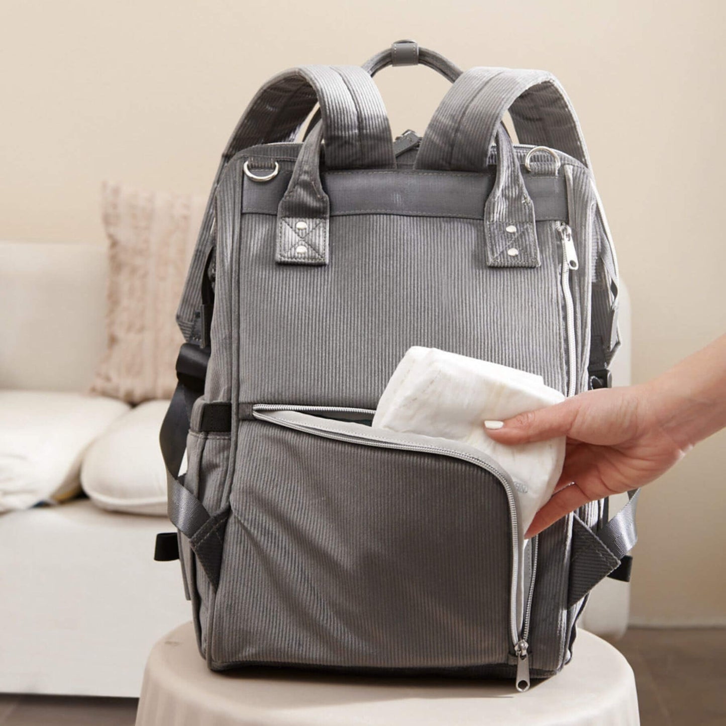 Corduroy Diaper Backpack Grey with a hand holding a white towel, placed on a beige surface.