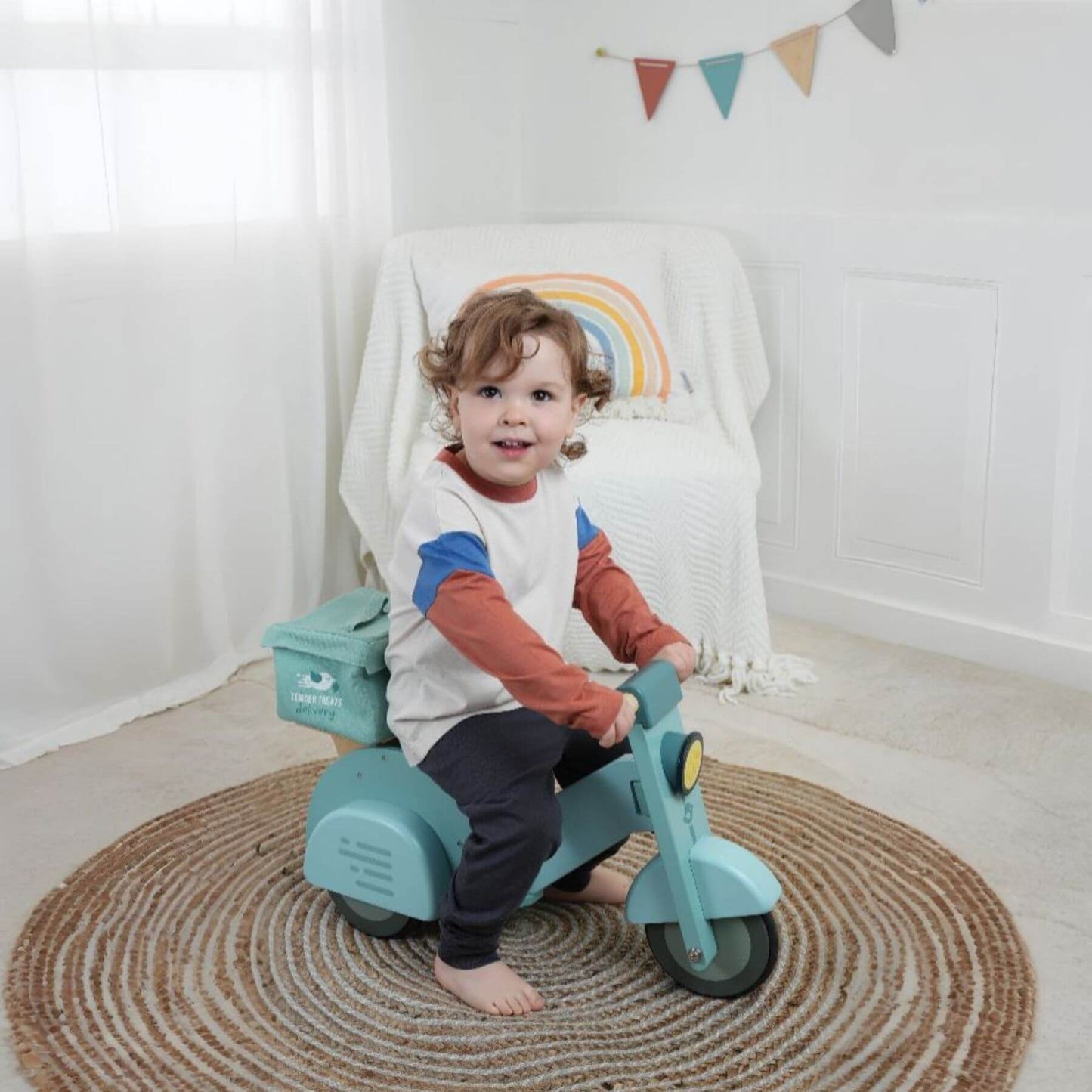 Child sitting on Tender Leaf Delivery Scooter in a room with a rainbow decoration.