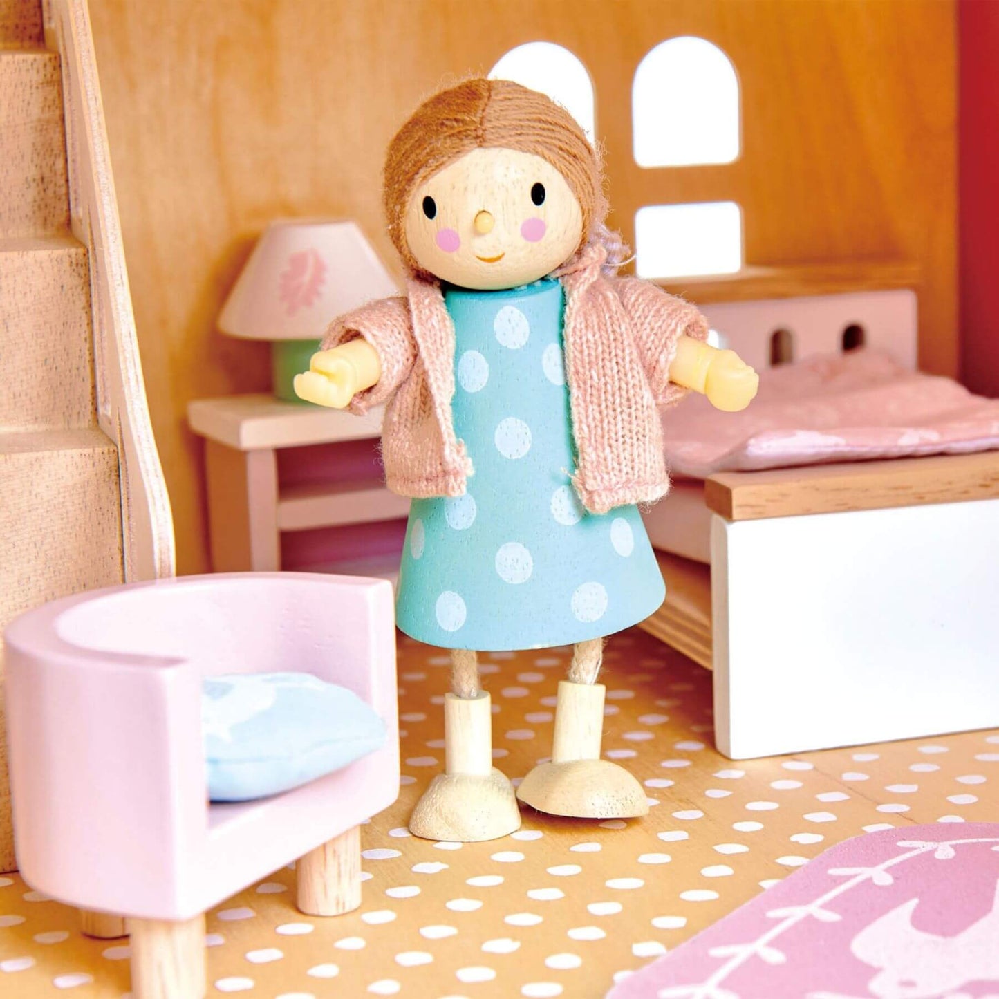 Wooden doll in a polka dot dress standing in a child's playroom with furniture.