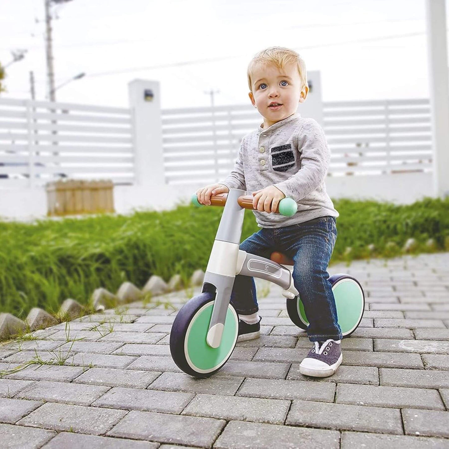 Child riding Hape First Ride Balance Bike Light Green on a paved path with grass and a white fence in the background