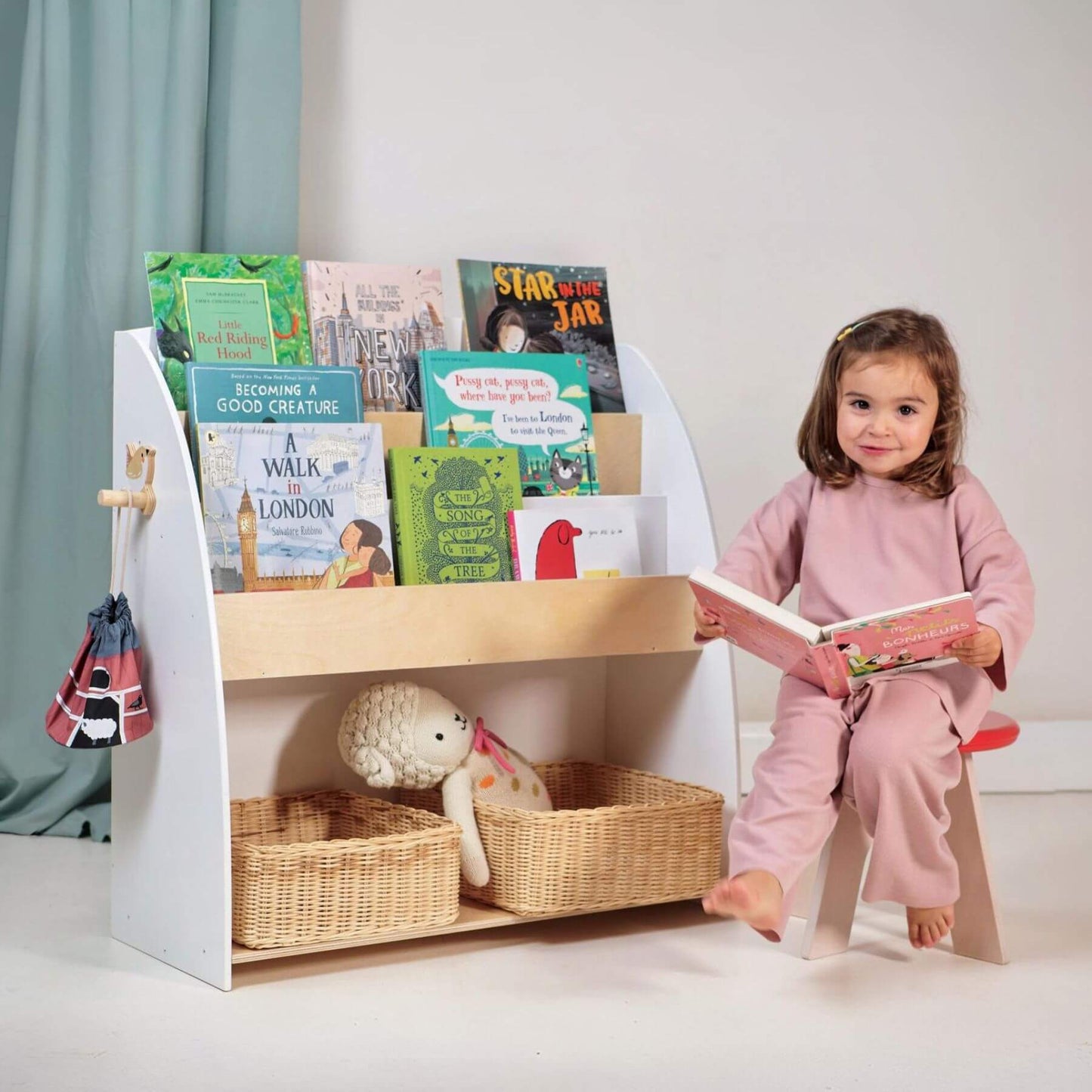 Child sitting on a stool reading a book next to Tender Leaf Forest Book Case with books and toys.