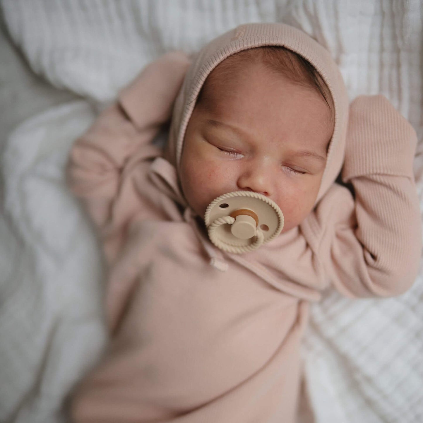 Newborn baby wrapped in a pink blanket with a FRIGG Pacifier, lying on a soft surface.