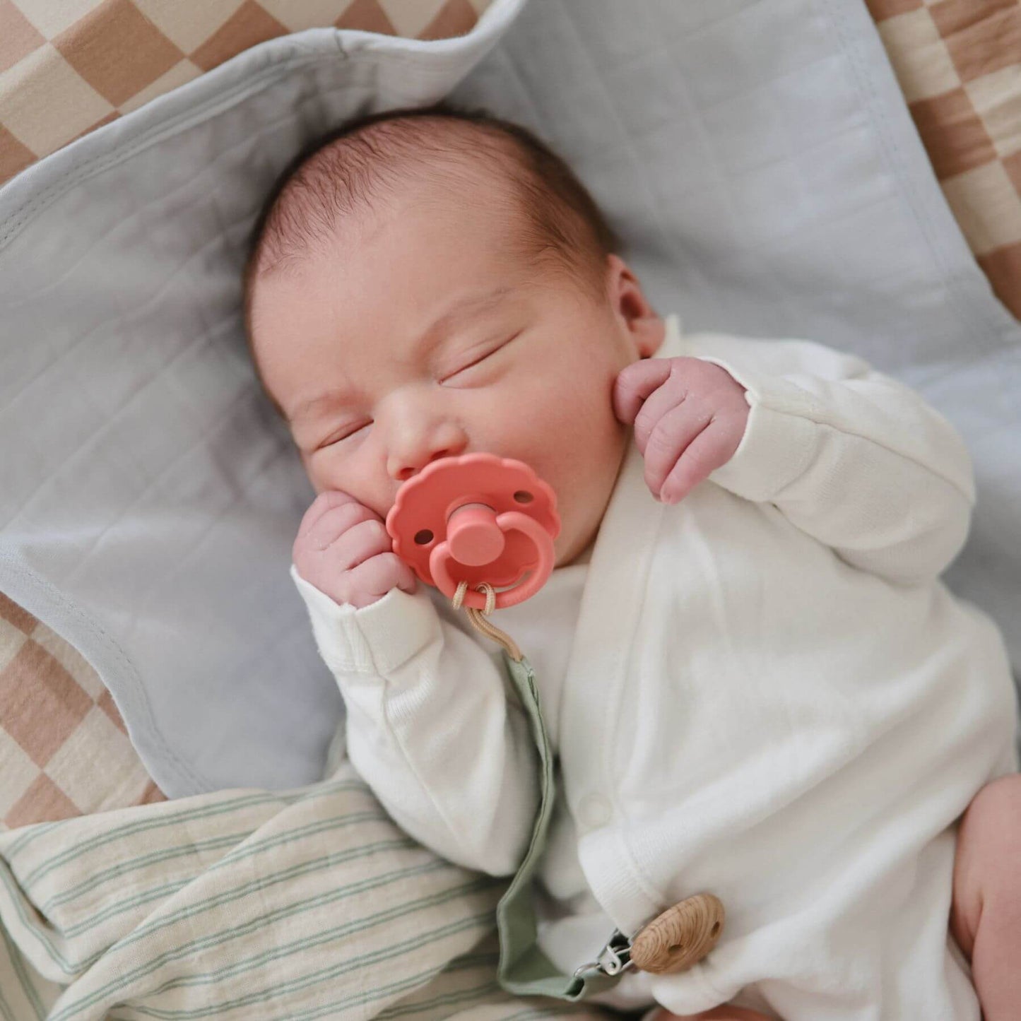 Newborn baby sleeping with a pink FRIGG pacifier in a soft, checkered crib.