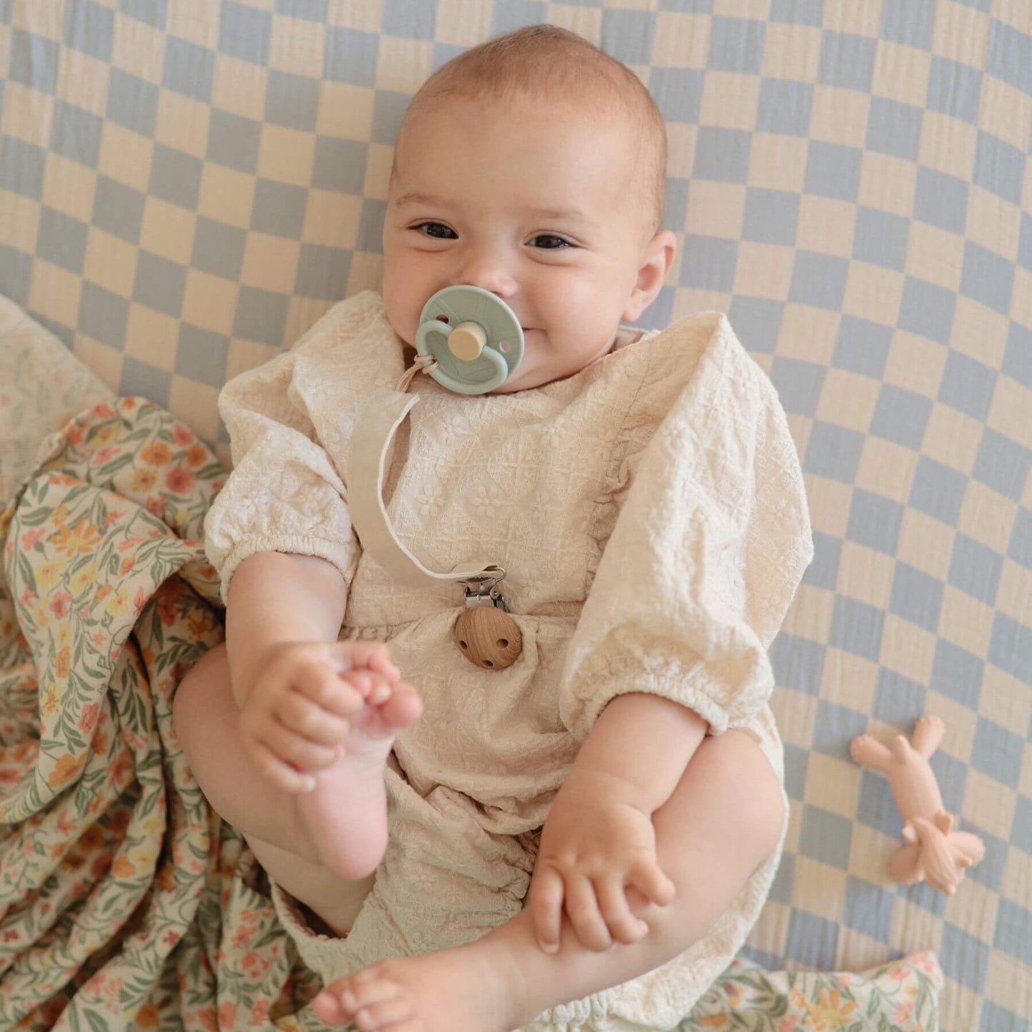 Baby in a beige outfit with a FRIGG pacifier, sitting on a checkered blanket.