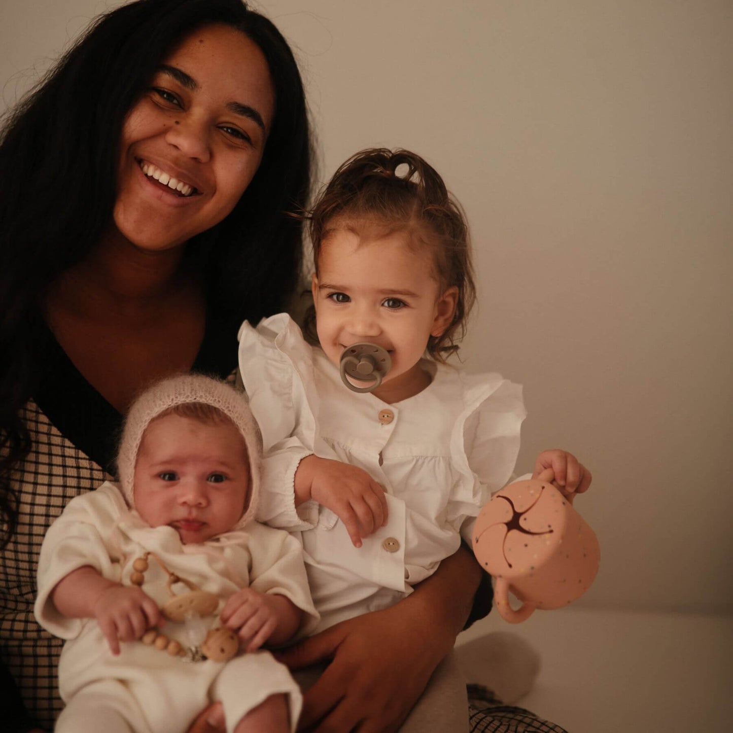 Woman holding two young children, one of whom is holding a toy, against a plain background.