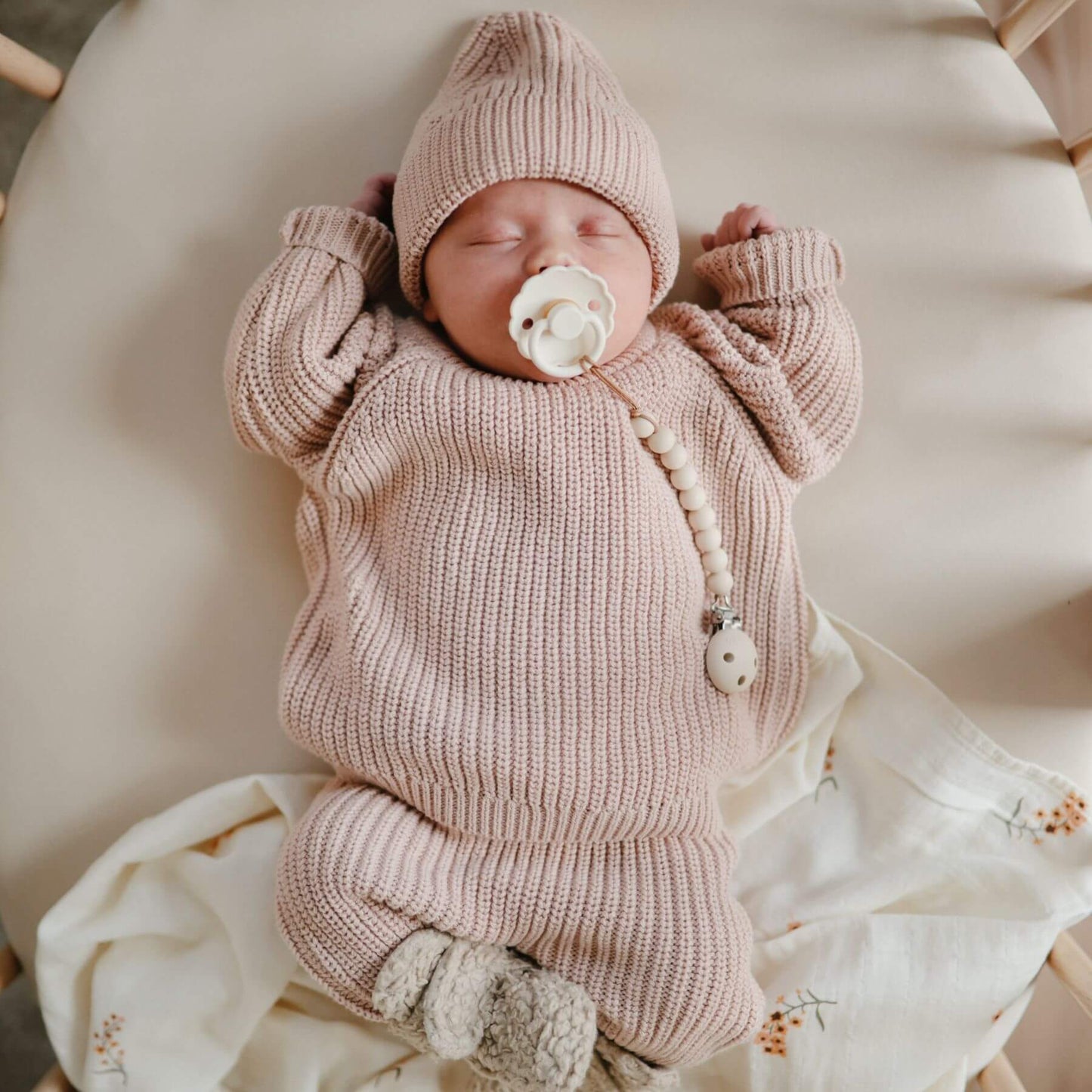 Newborn baby in a pink knitted outfit with a matching hat, FRIGG pacifier, lying on a soft surface.