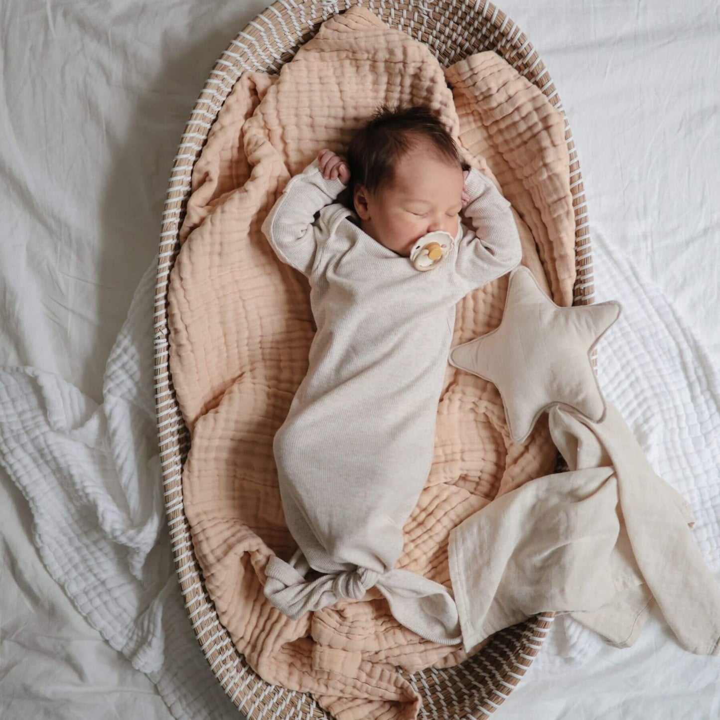 Newborn baby wrapped in a white blanket with a FRIGG pacifier, lying in a wicker basket with star-shaped pillow.