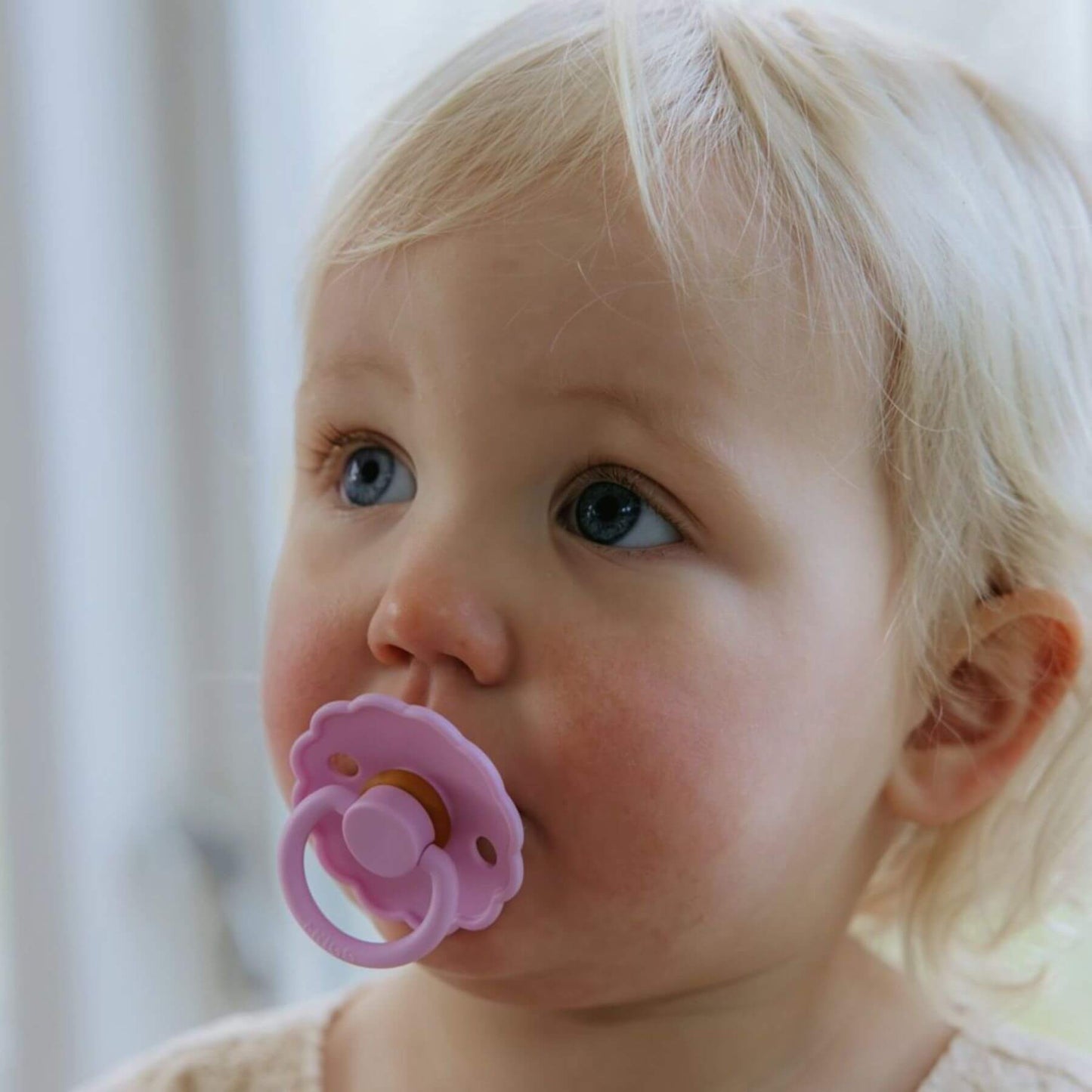 Baby with a pink FRIGG pacifier in a blurred indoor setting