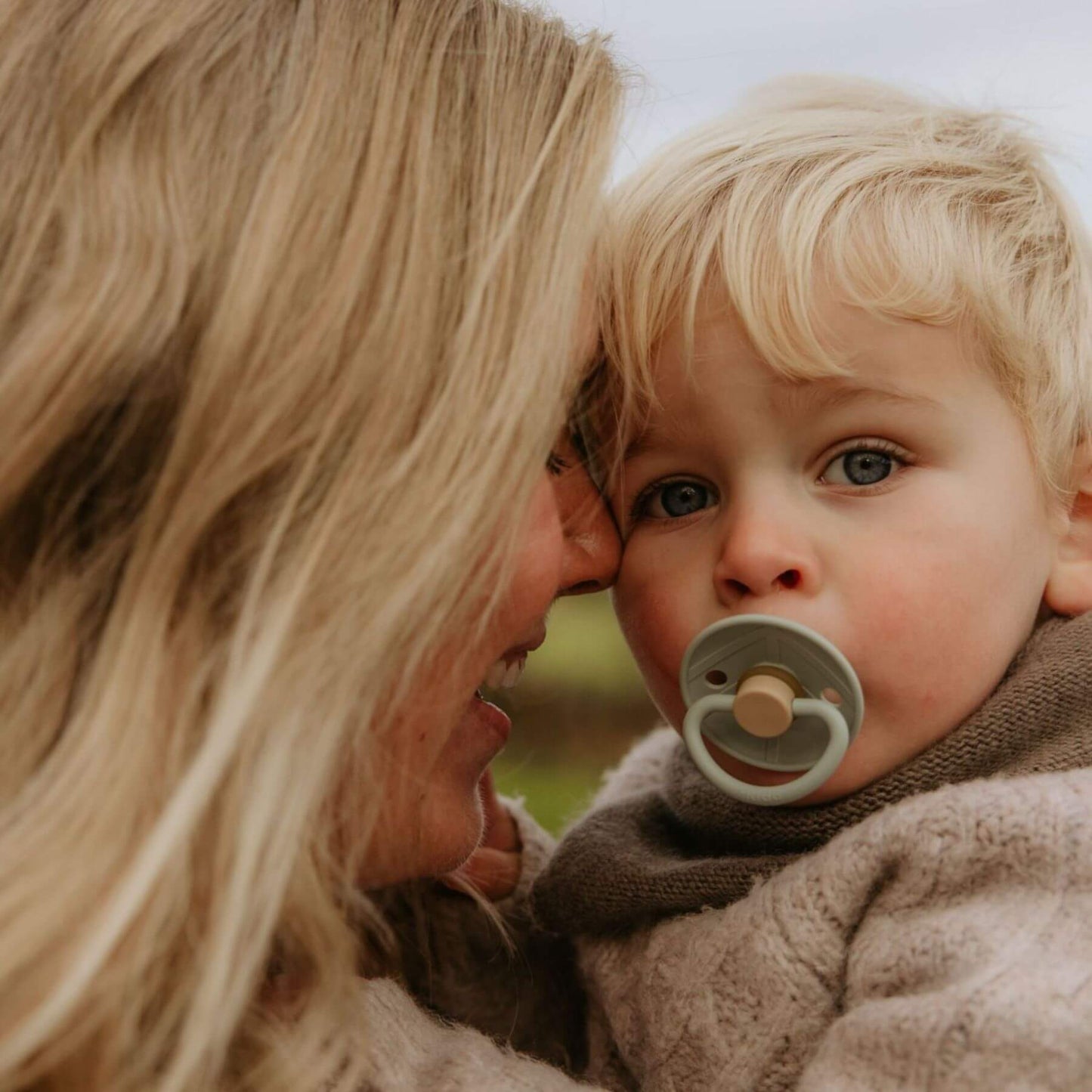 Woman holding a child with a FRIGG pacifier, both looking at each other outdoors.