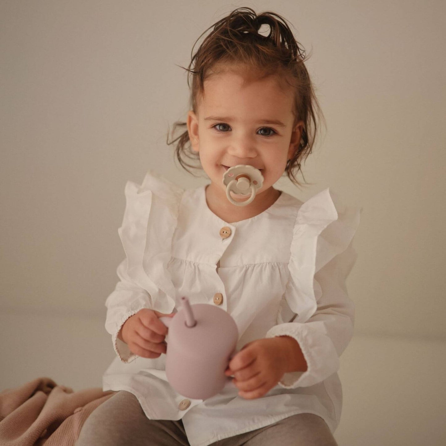 Child holding a pink toy, holding a FRIGG pacifier in her mouth
