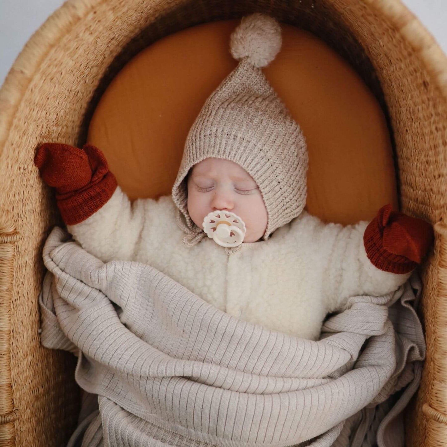 Newborn baby sleeping in a woven basket with a knitted hat and FRIGG pacifier.