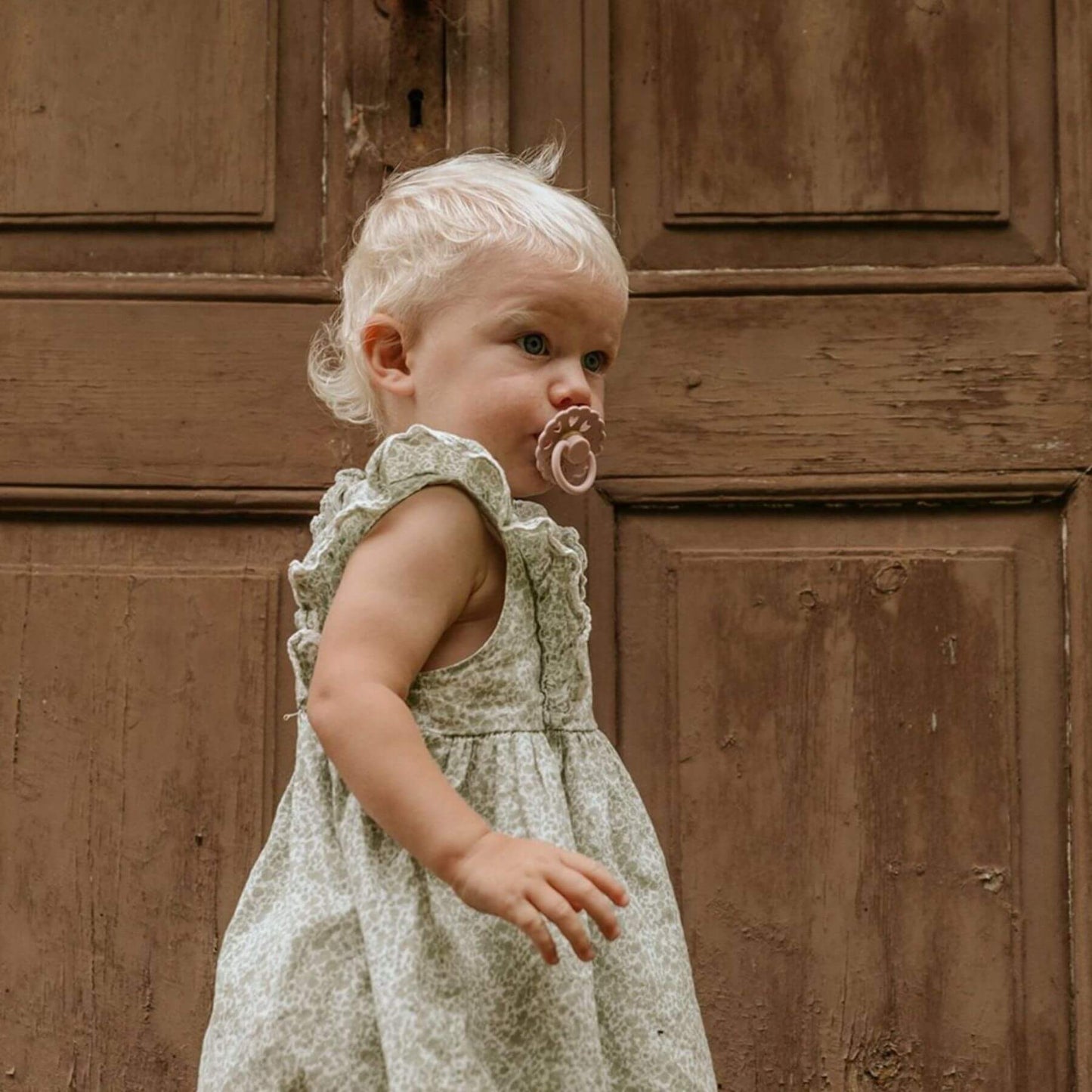 Child in a floral dress with a FRIGG pacifier against a wooden door background