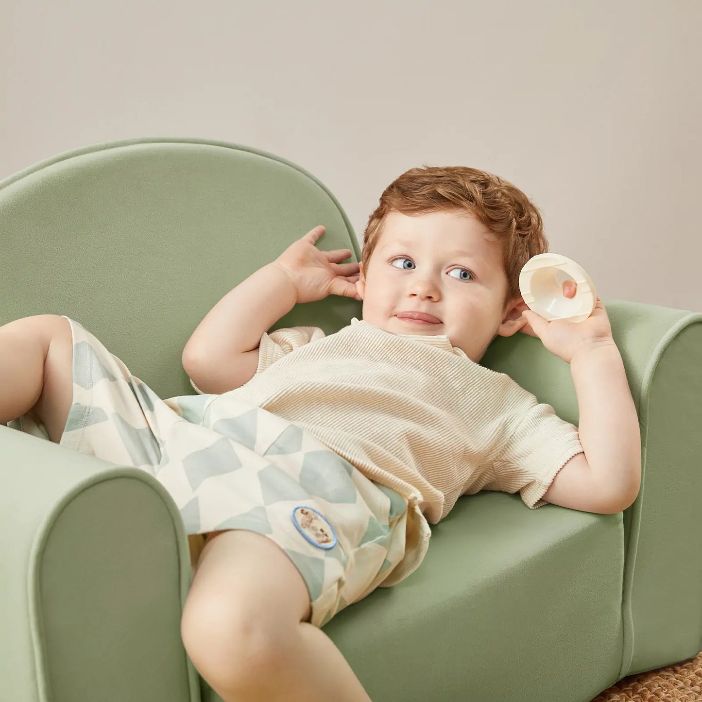 Child sitting on Tiny Land® GentleSnug Kid Chair Sage holding a small object, wearing a light-colored outfit with patterns.