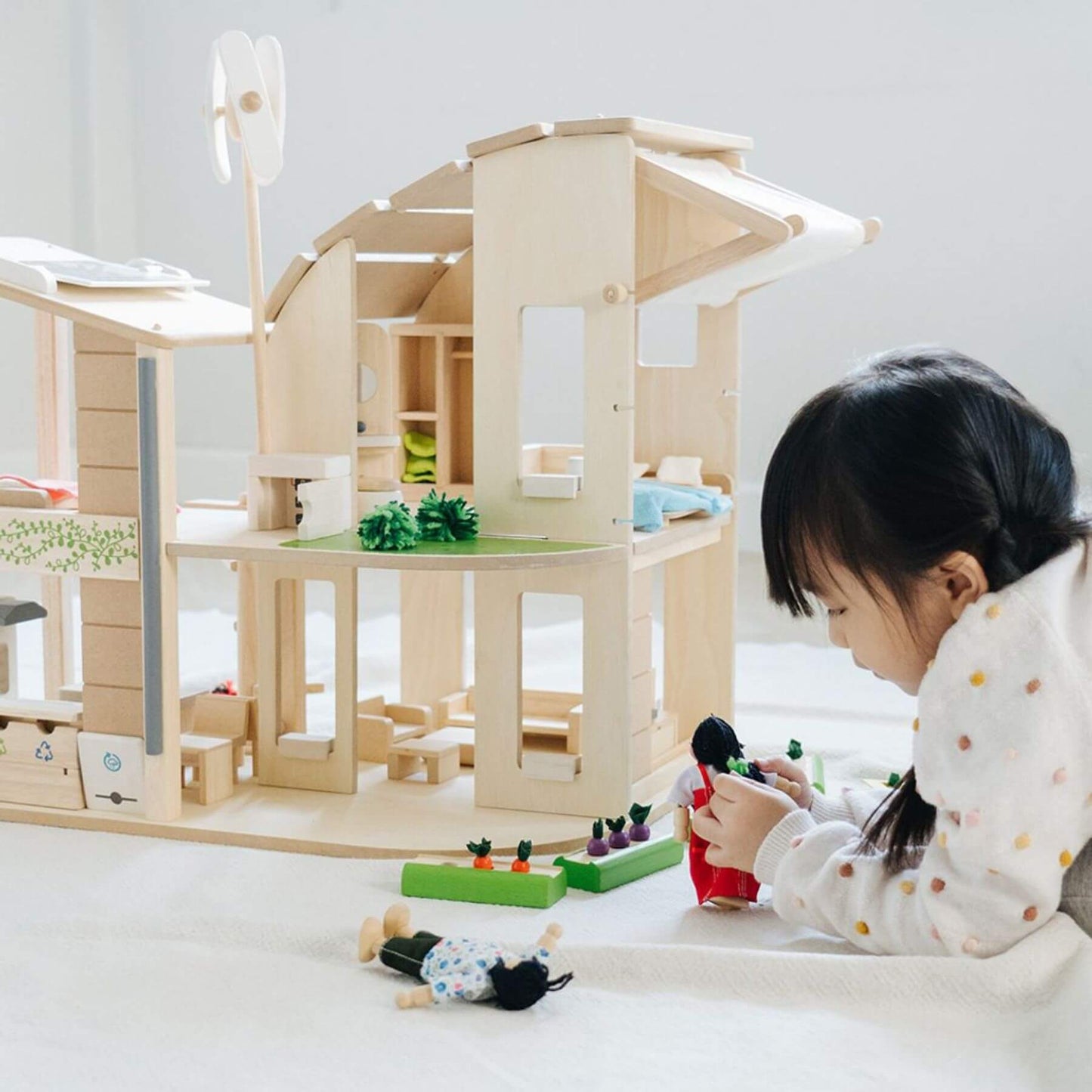 Child playing with PlanToys Green Dollhouse With Furniture and toys on a light-colored floor.