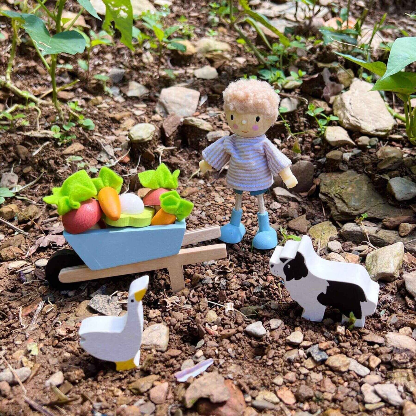 Toy figure with a wheelbarrow full of vegetables, a toy cow, and a toy duck on a rocky ground.