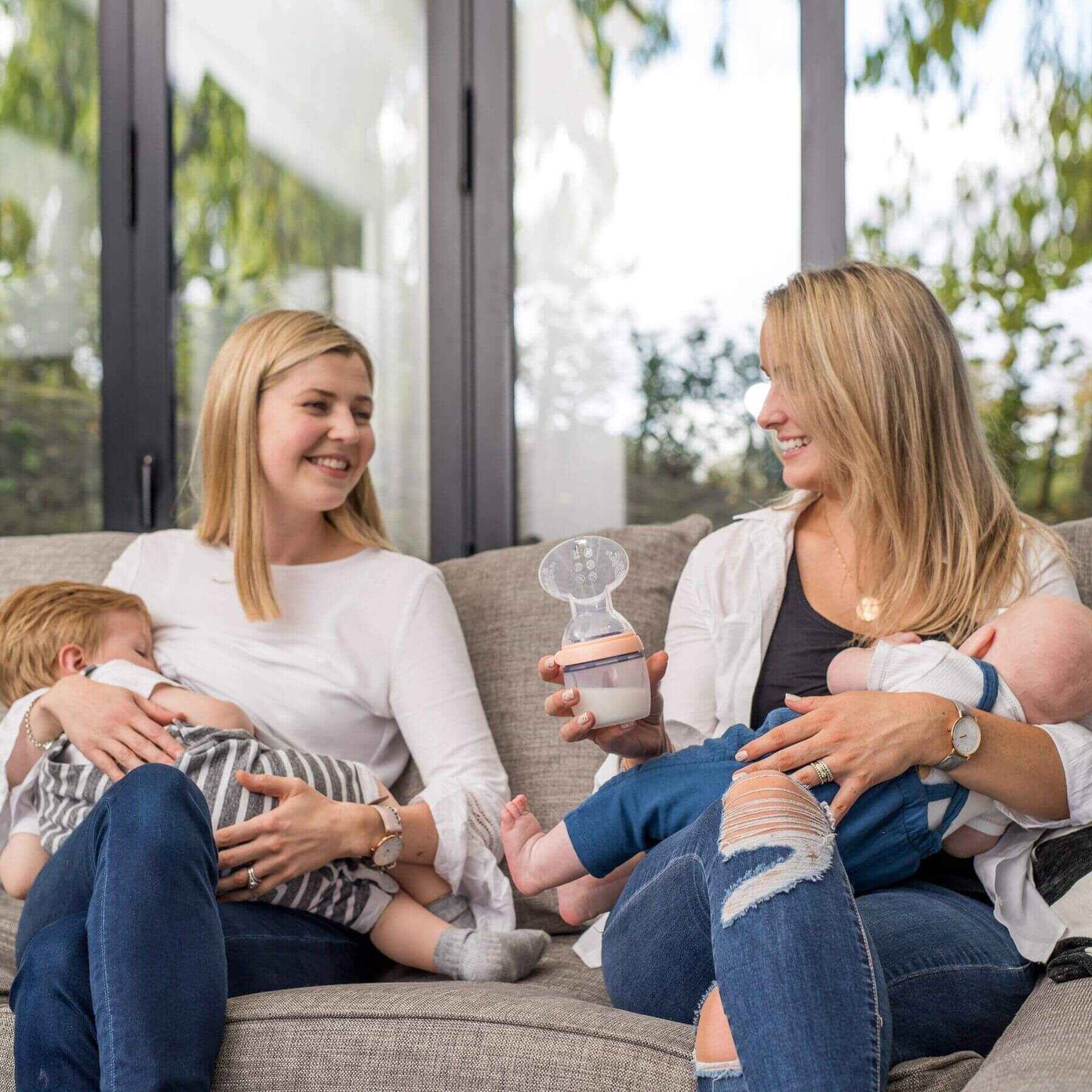 Two women sitting on a couch with babies, one holding a breast pump.
