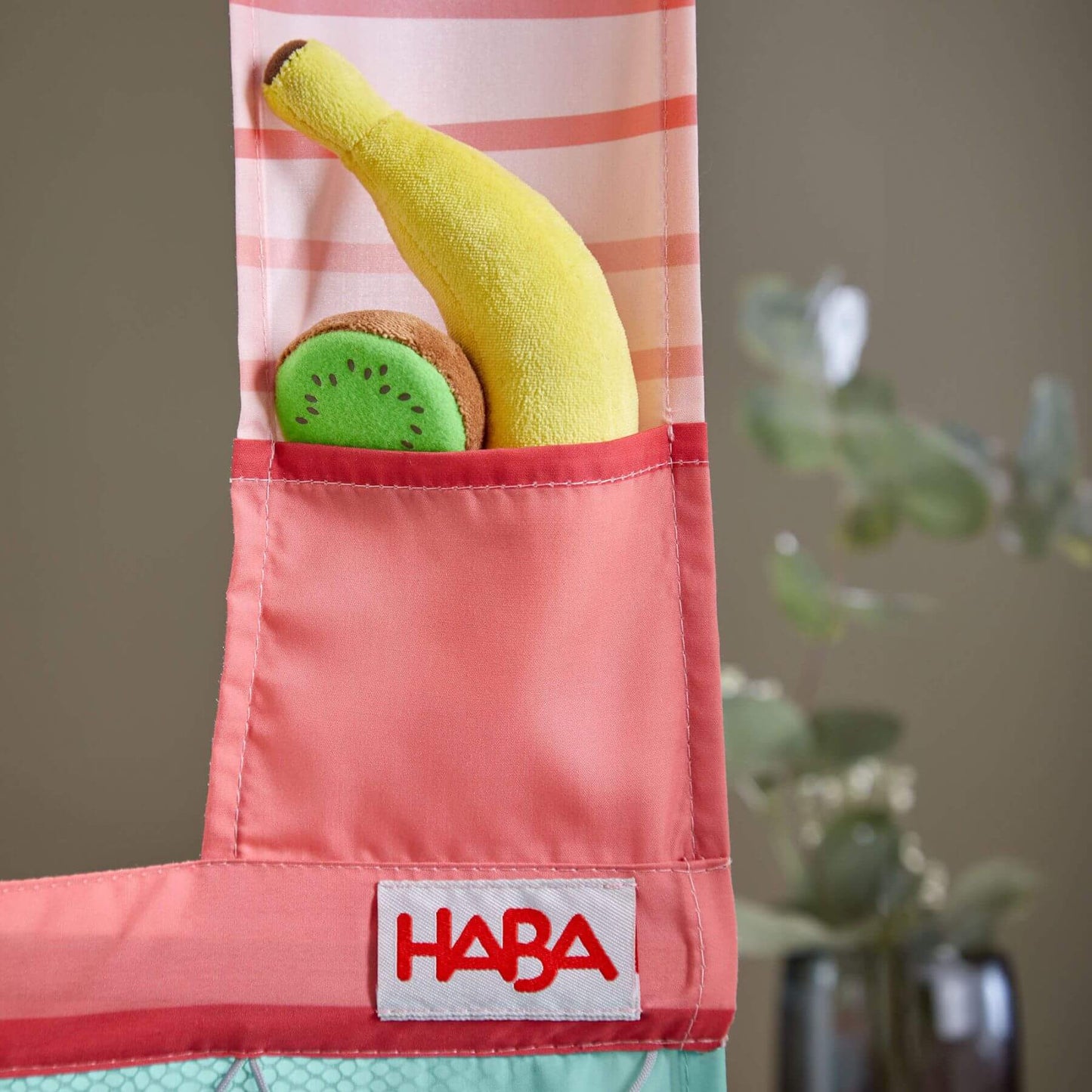 Pink toy apron with banana and kiwi plush toys, HABA logo visible, plant in the background