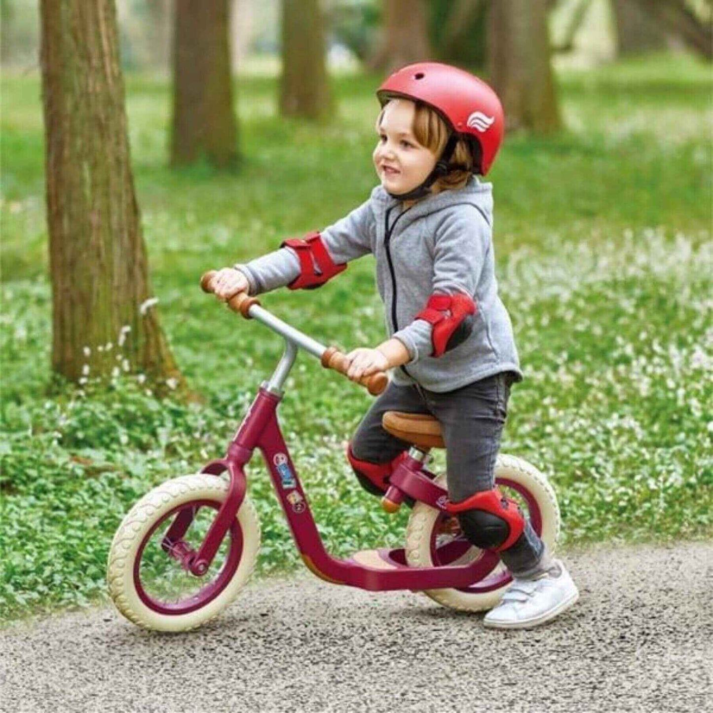 Child riding Hape Learn to Ride Balance Bike Red with safety gear in a park setting