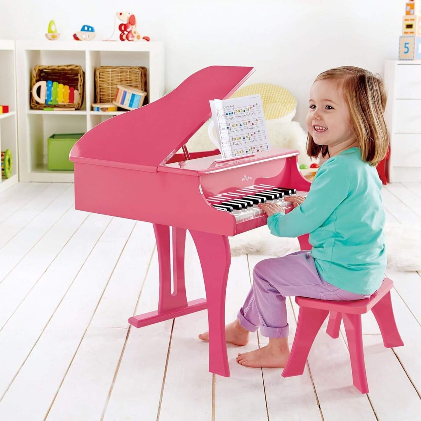 Child playing Hape Happy Grand Piano Pink in a bright room with white floor and shelves.