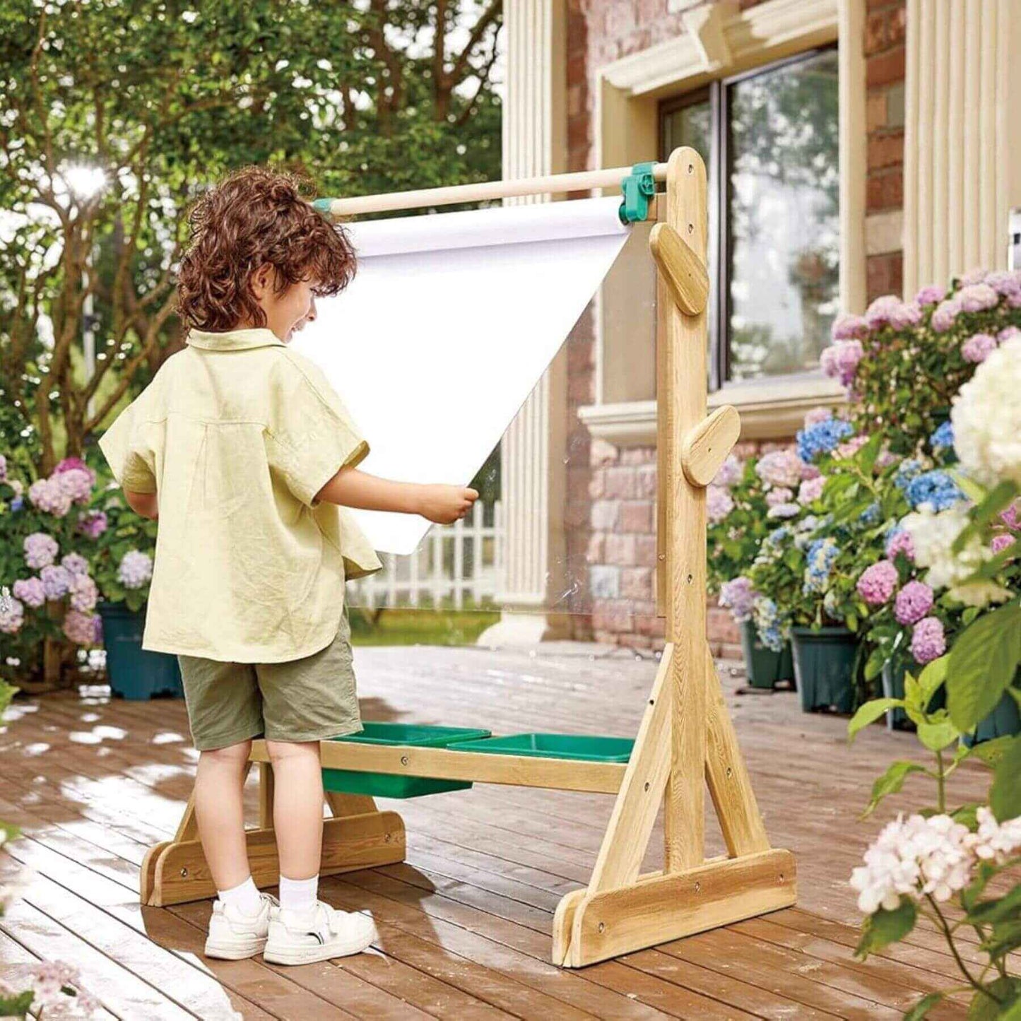 Child using Hape Outdoor Art Easel with flowers and a house in the background